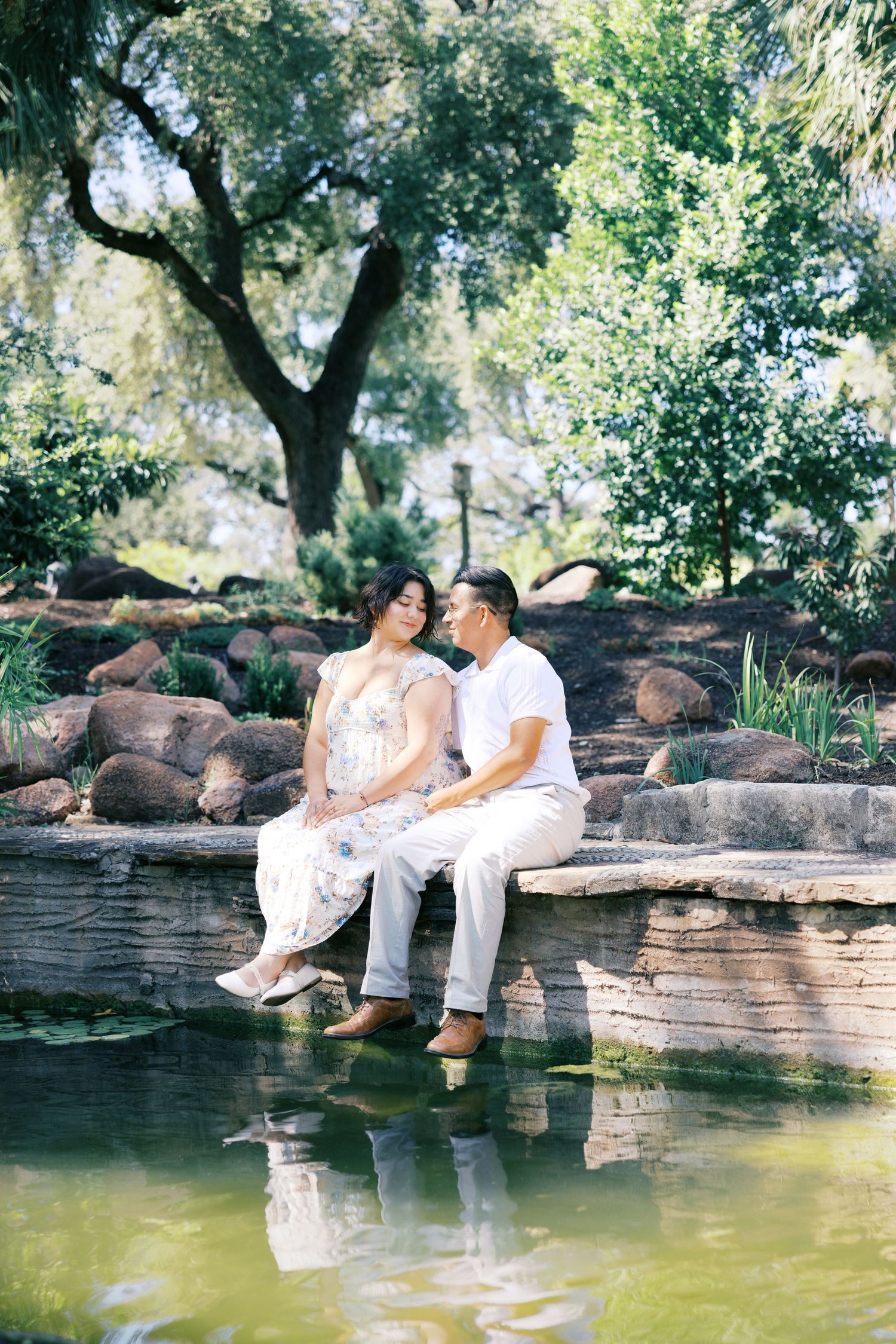 A couple sits together on a stone ledge by a tranquil pond in a garden setting.