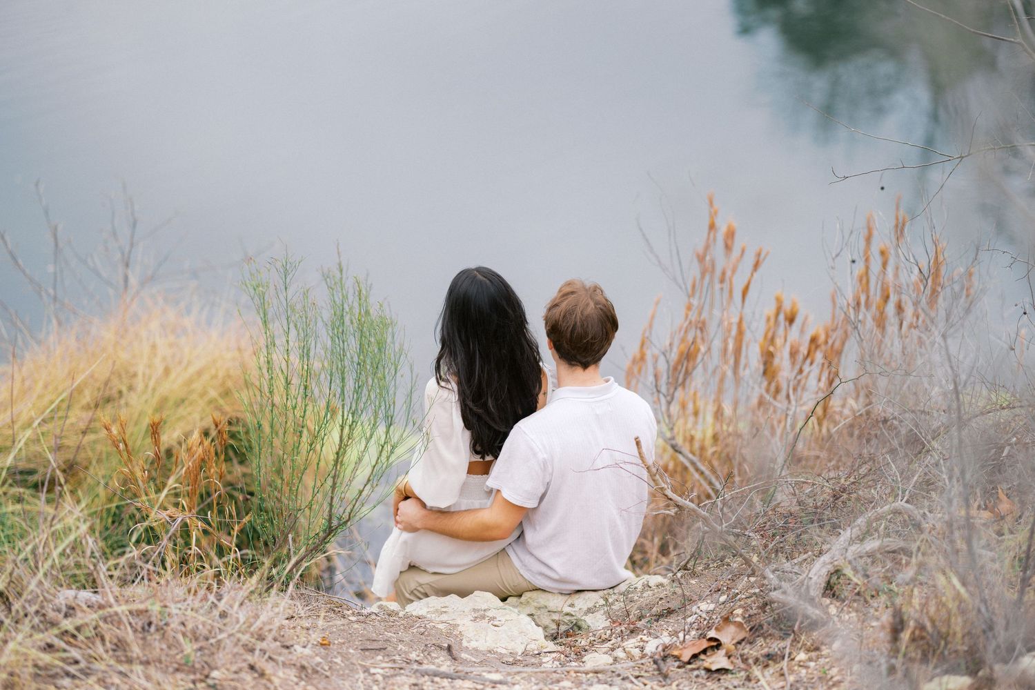 Two people sitting close together on lakeside rocks during golden hour.