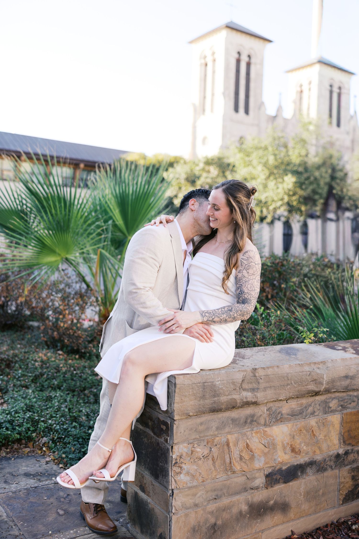 Romantic couple sitting on stone wall with church and palm trees in background.