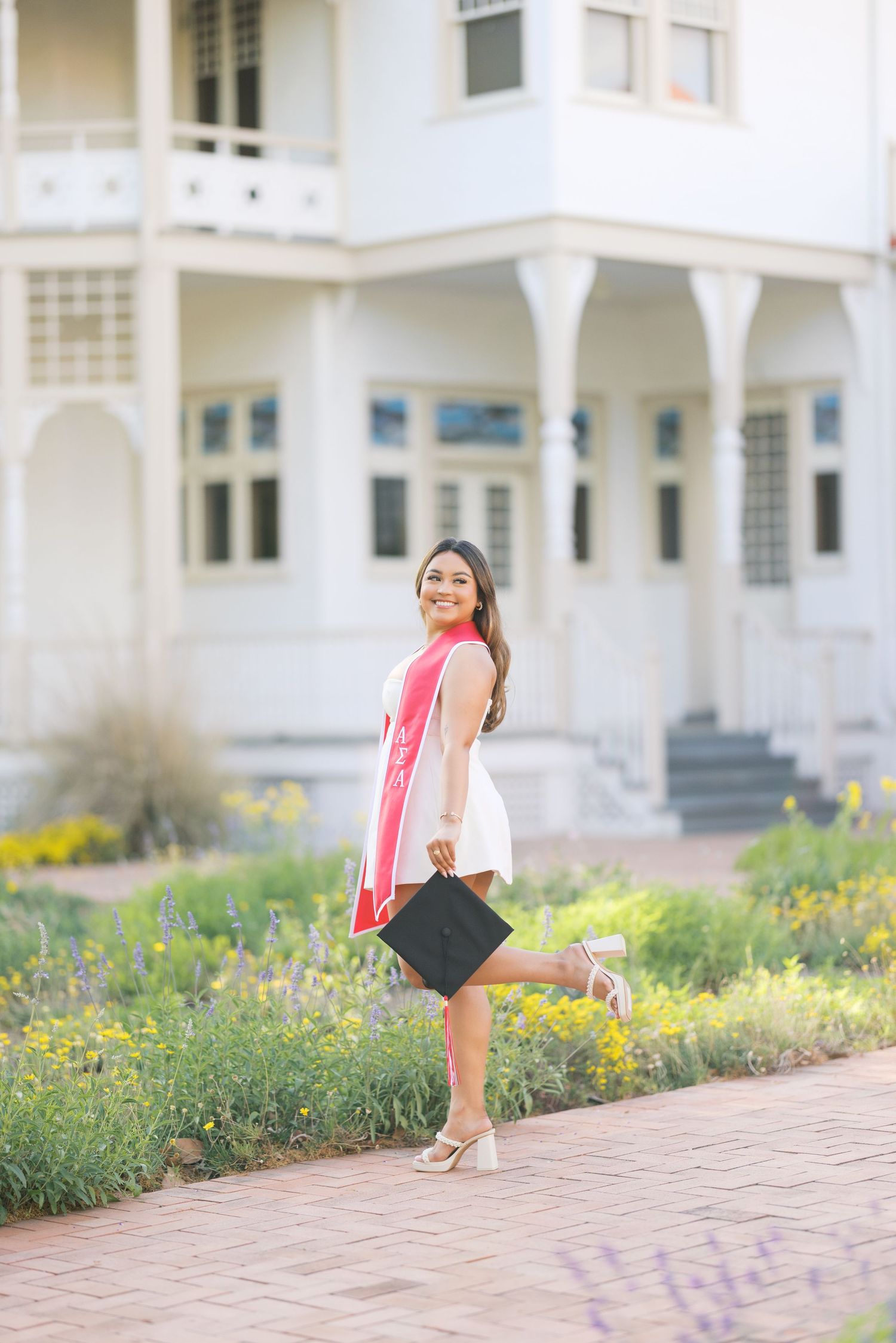 Smiling graduate in white dress and red sash holds cap on brick path outside white building.