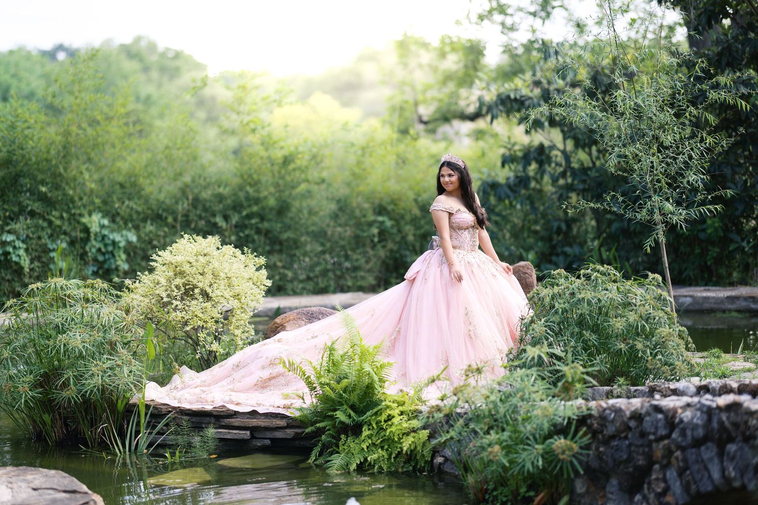 Young woman in elegant pink ball gown poses on stone bridge surrounded by lush green garden and tranquil pond.