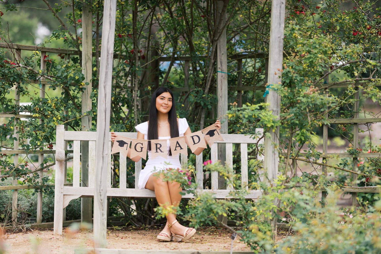 Young woman sitting on a white garden bench holding a GRAD banner, surrounded by lush greenery for graduation photos.
