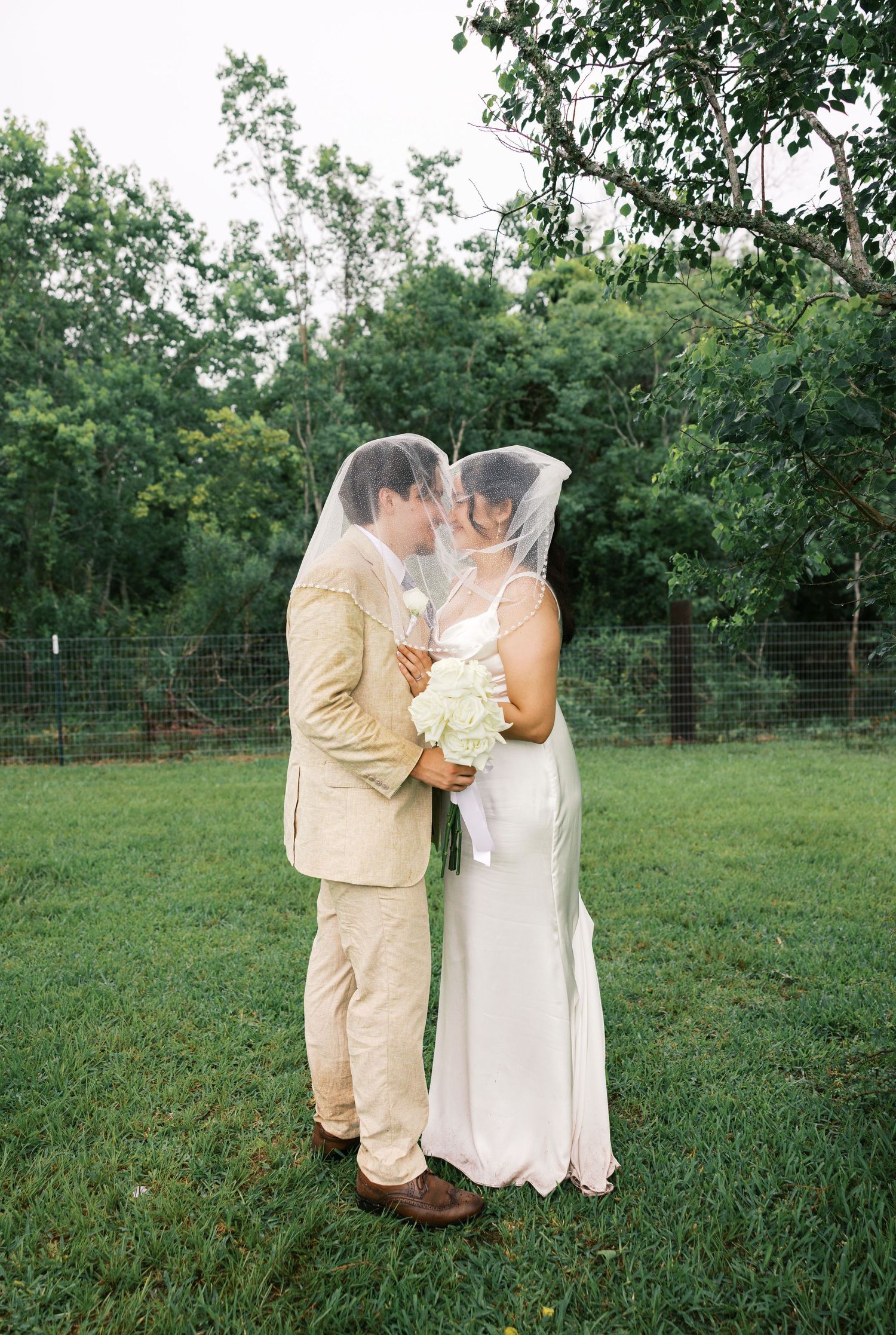 Bride and groom sharing a romantic kiss under a veil outdoors, surrounded by lush green trees.