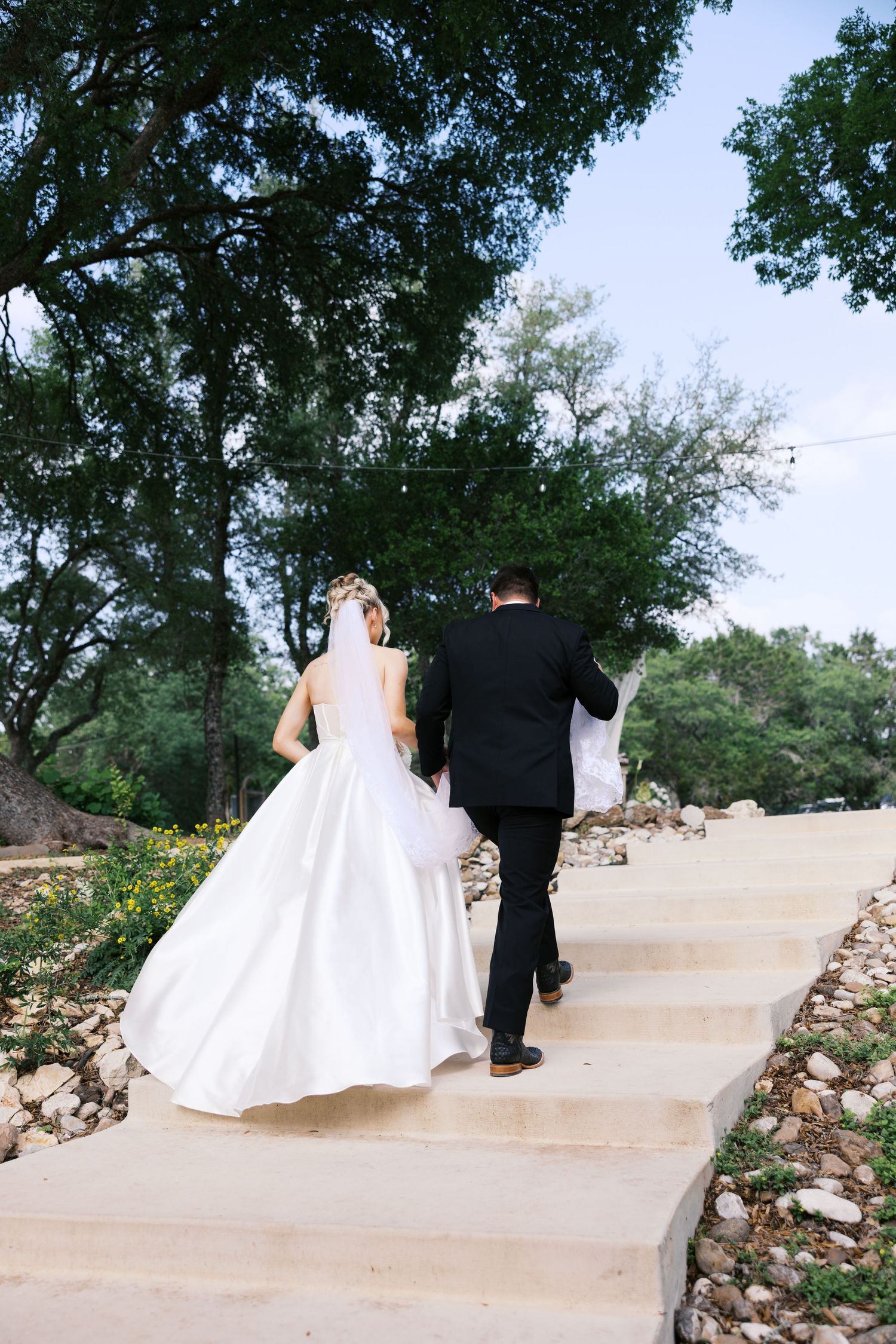Bride in white gown and groom in black tuxedo walk up stone steps surrounded by lush green trees.