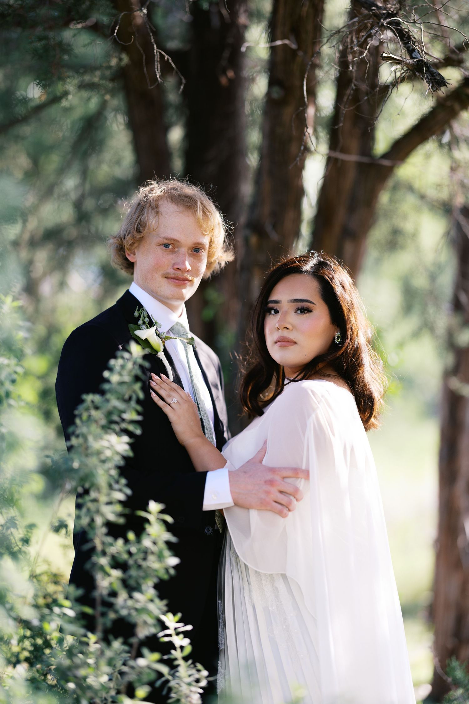 A couple poses in a sunlit forest, the groom in a black tuxedo and the bride in a flowing white gown.