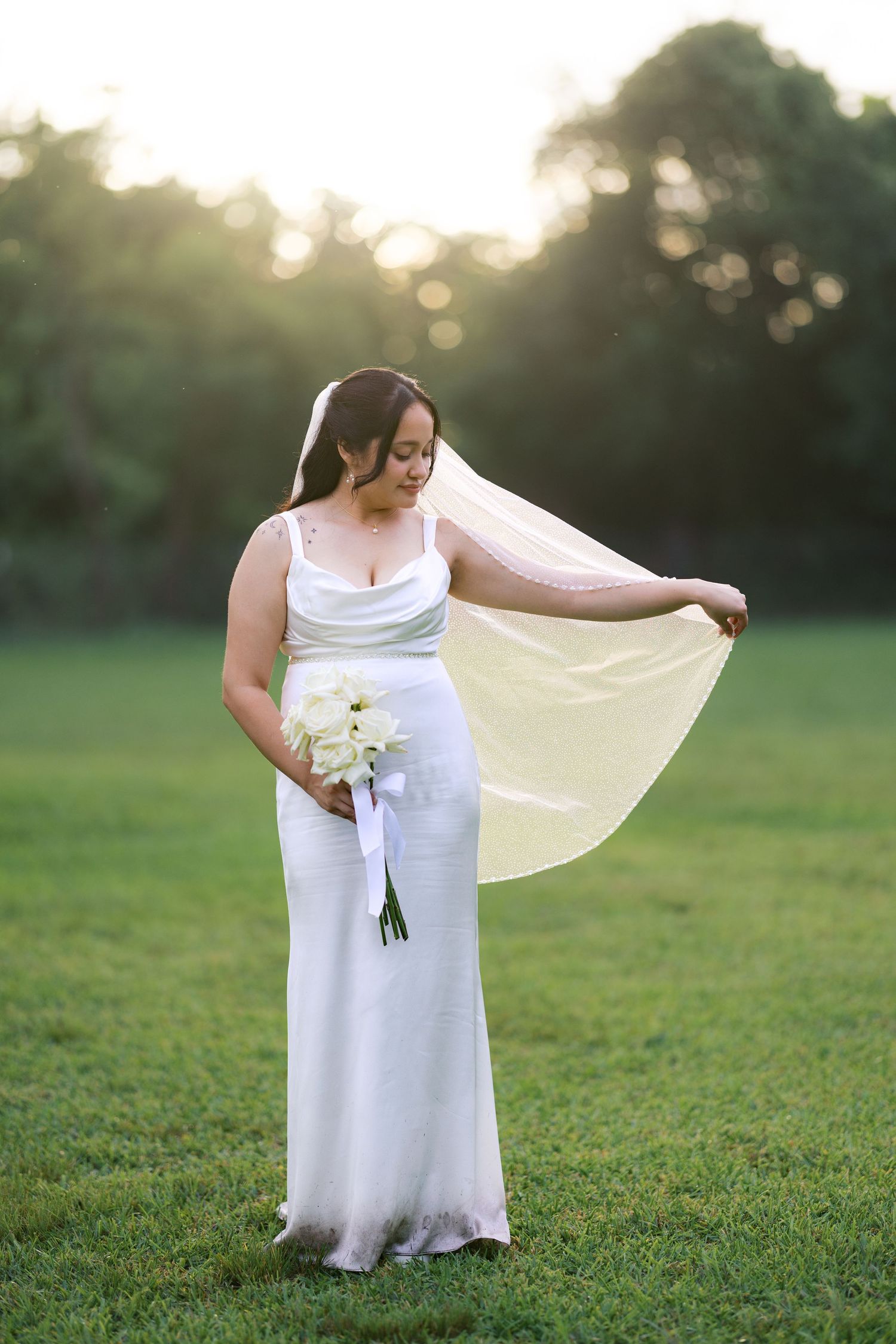 Bride in white gown holds bouquet while lifting veil in sunlit green field at golden hour.