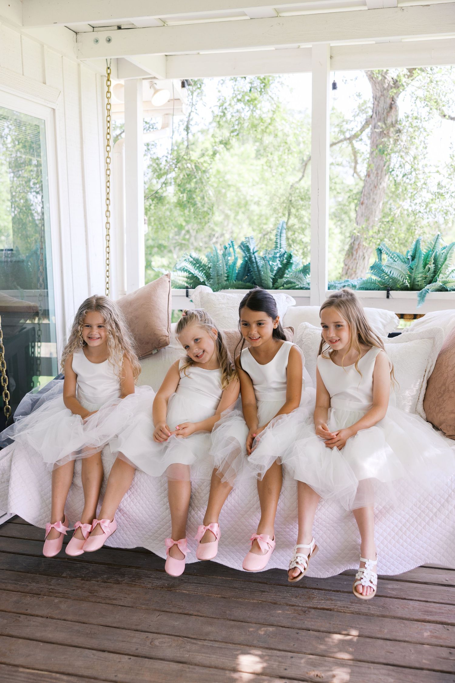 Four flower girls in white tulle dresses and pink shoes sit together smiling on a porch swing.