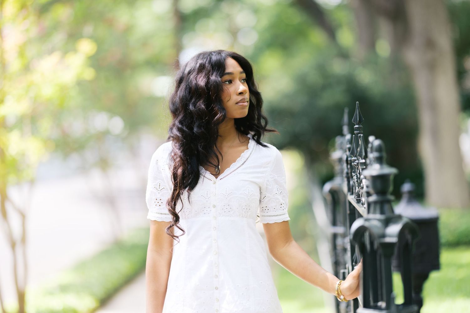 Young Black woman in white lace dress poses gracefully by ornate iron gate surrounded by lush green trees.