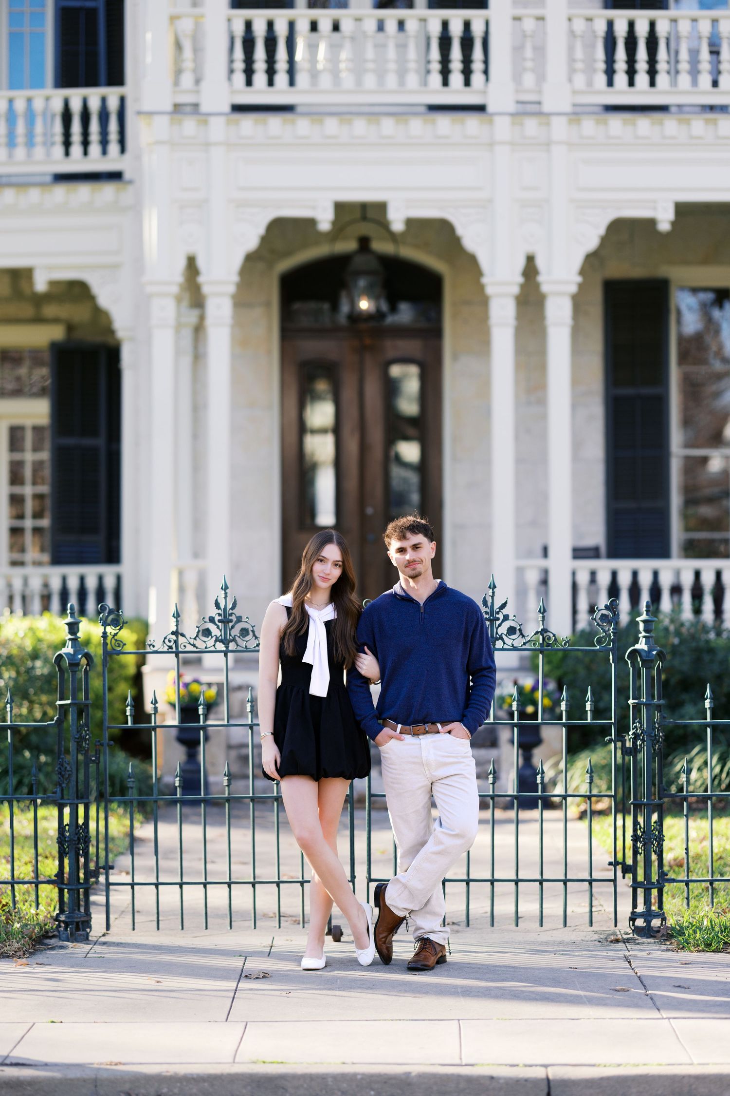 A young couple poses in front of a grand white Southern mansion with wrought iron fence.