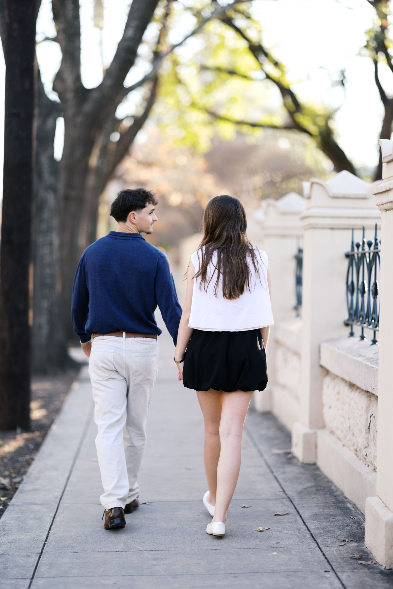 Couple walking hand in hand down a sunny sidewalk, man in navy sweater and woman in black skirt seen from behind.
