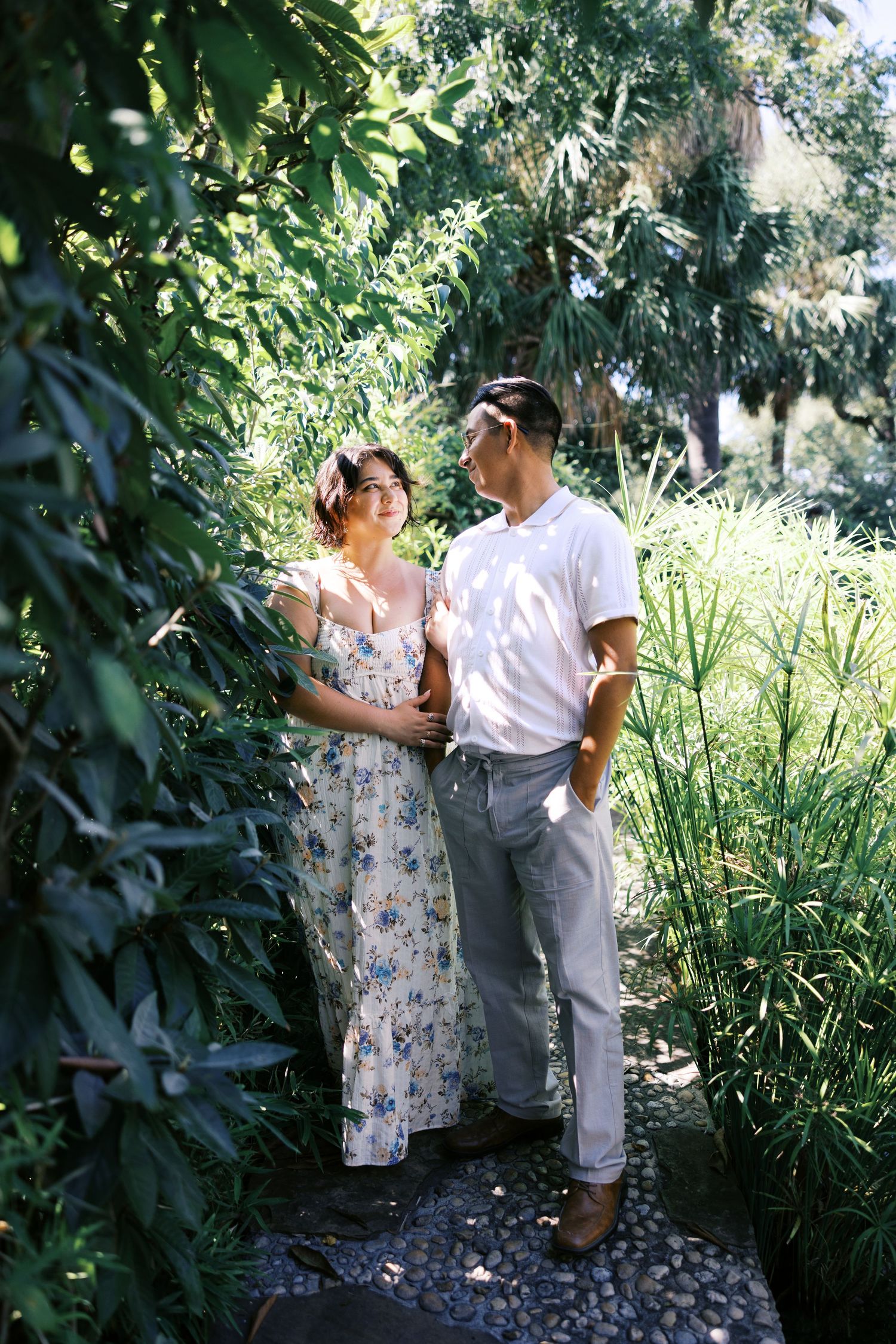 A couple poses together on a garden path surrounded by lush greenery and natural light.
