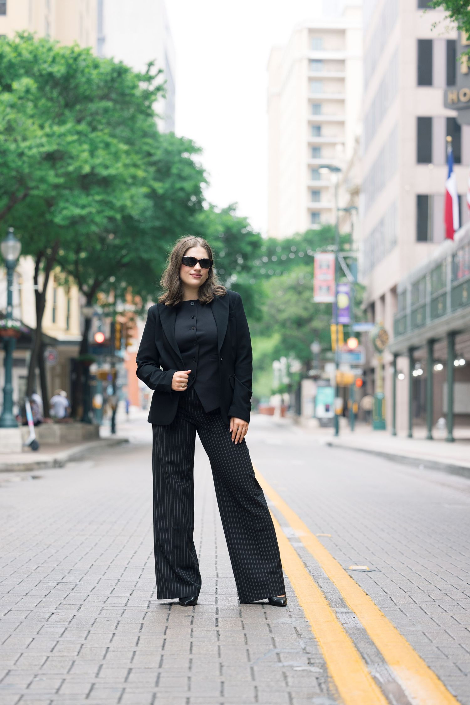 Person in black attire and sunglasses posing on a city street with yellow road markings.
