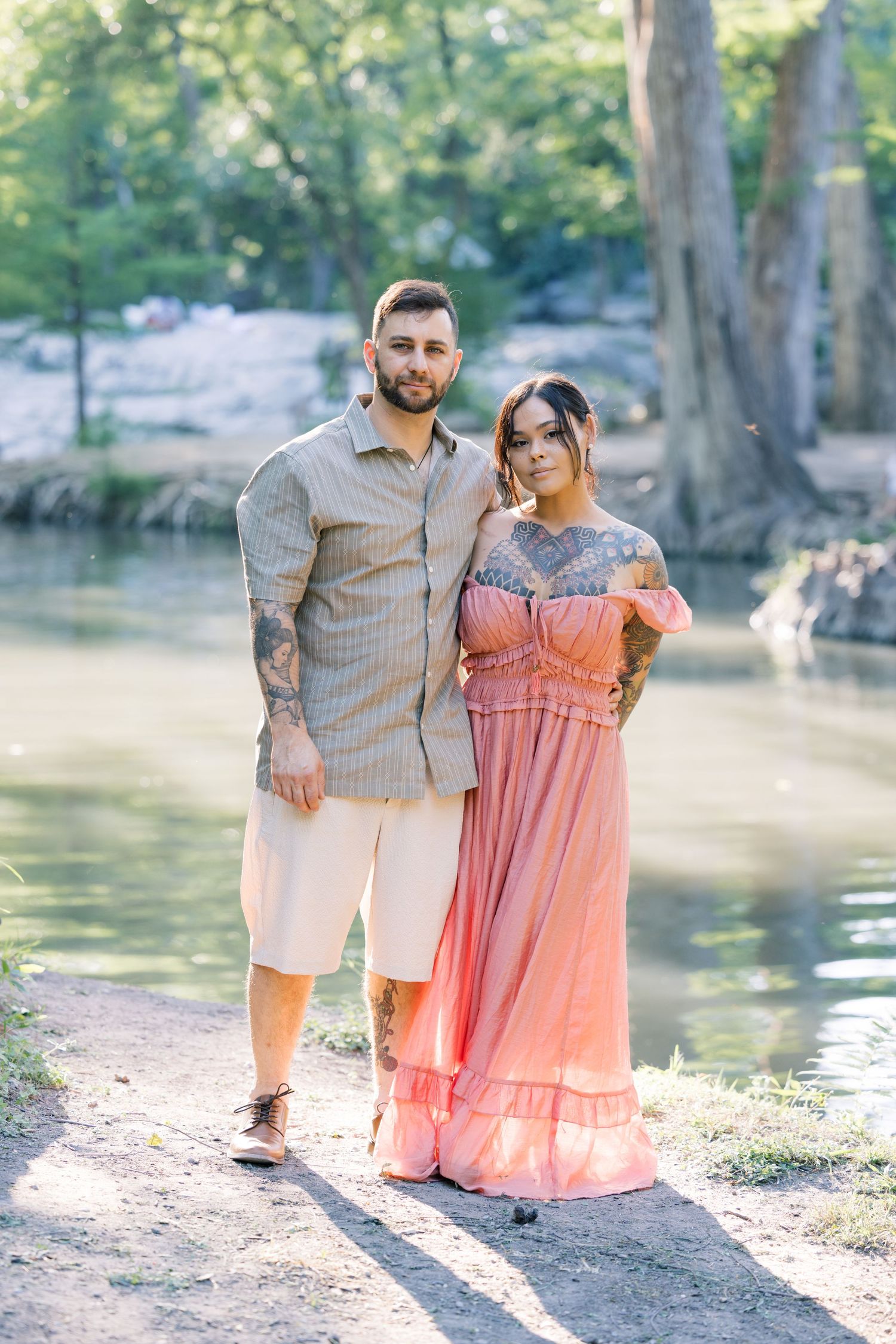 A couple poses by a lake in coordinating coral and beige outfits during a sunny outdoor photoshoot.