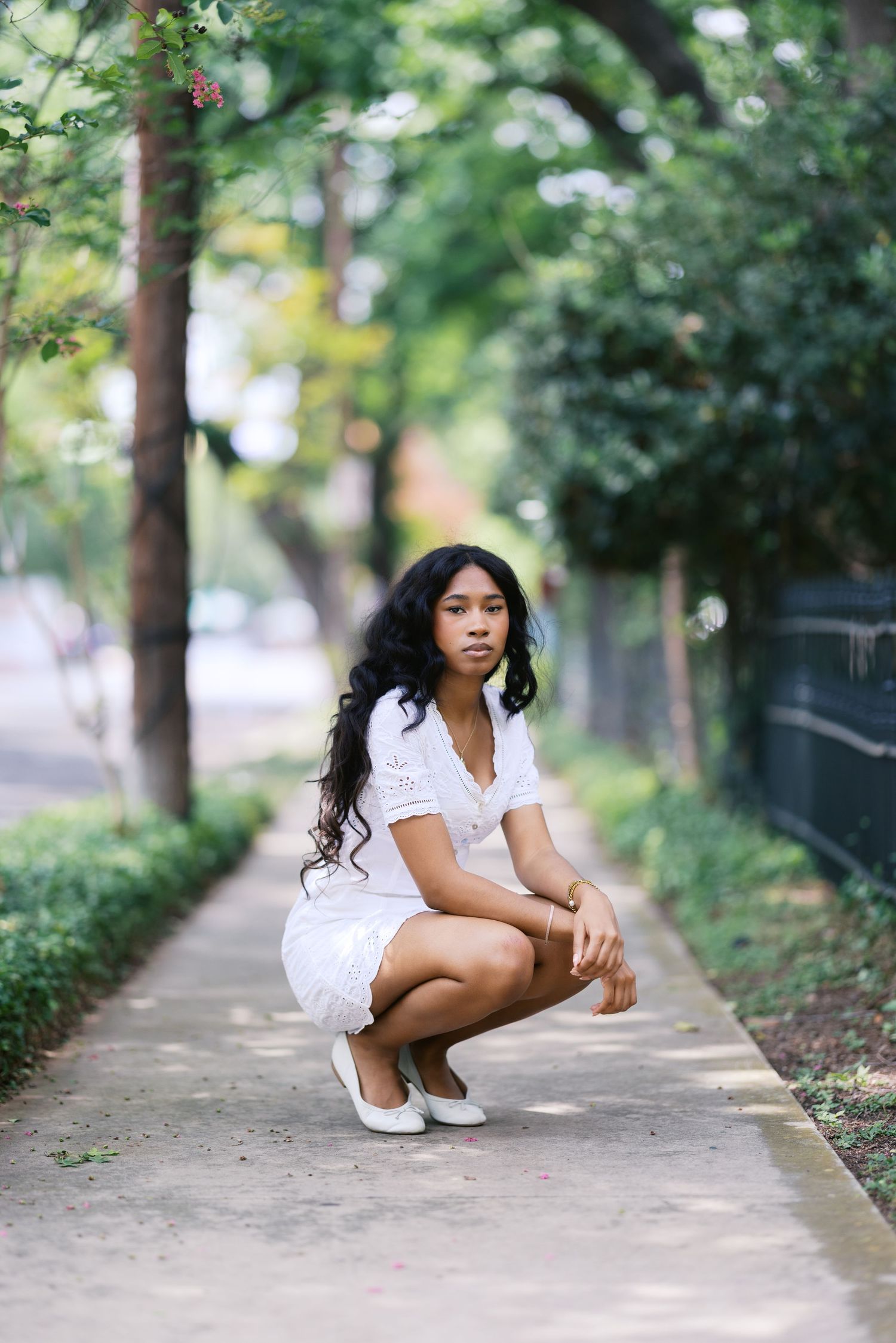 Young Black woman in white lace dress crouching on a tree-lined sidewalk path.