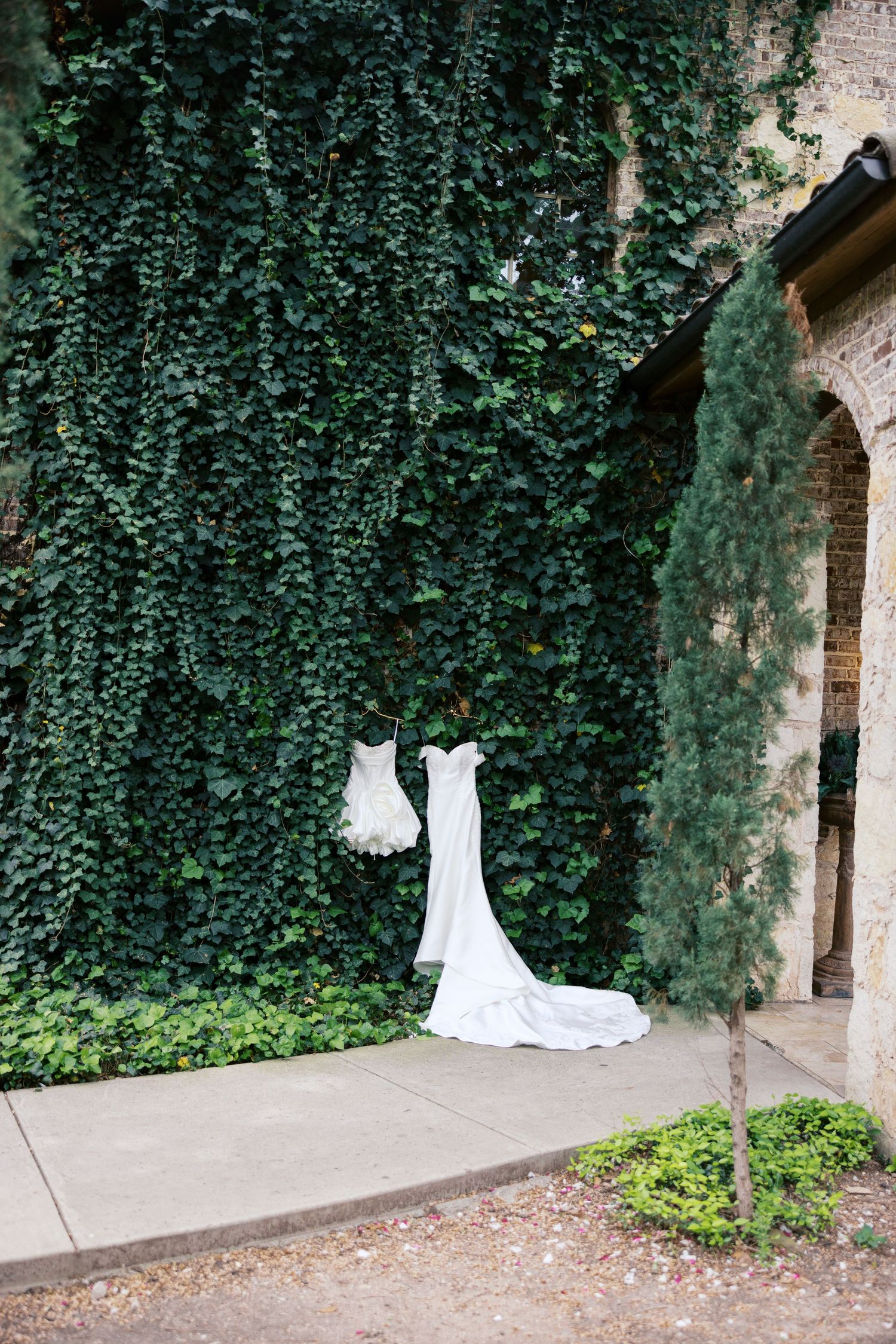 White wedding dress and veil hanging on ivy-covered stone wall at elegant outdoor venue.