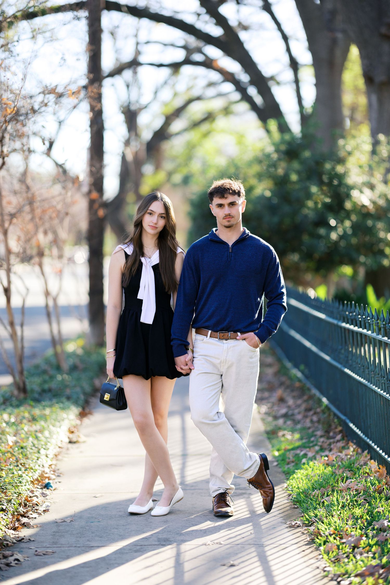 Young couple walking hand in hand on a sunny park path, woman in black dress, man in navy sweater and khakis.