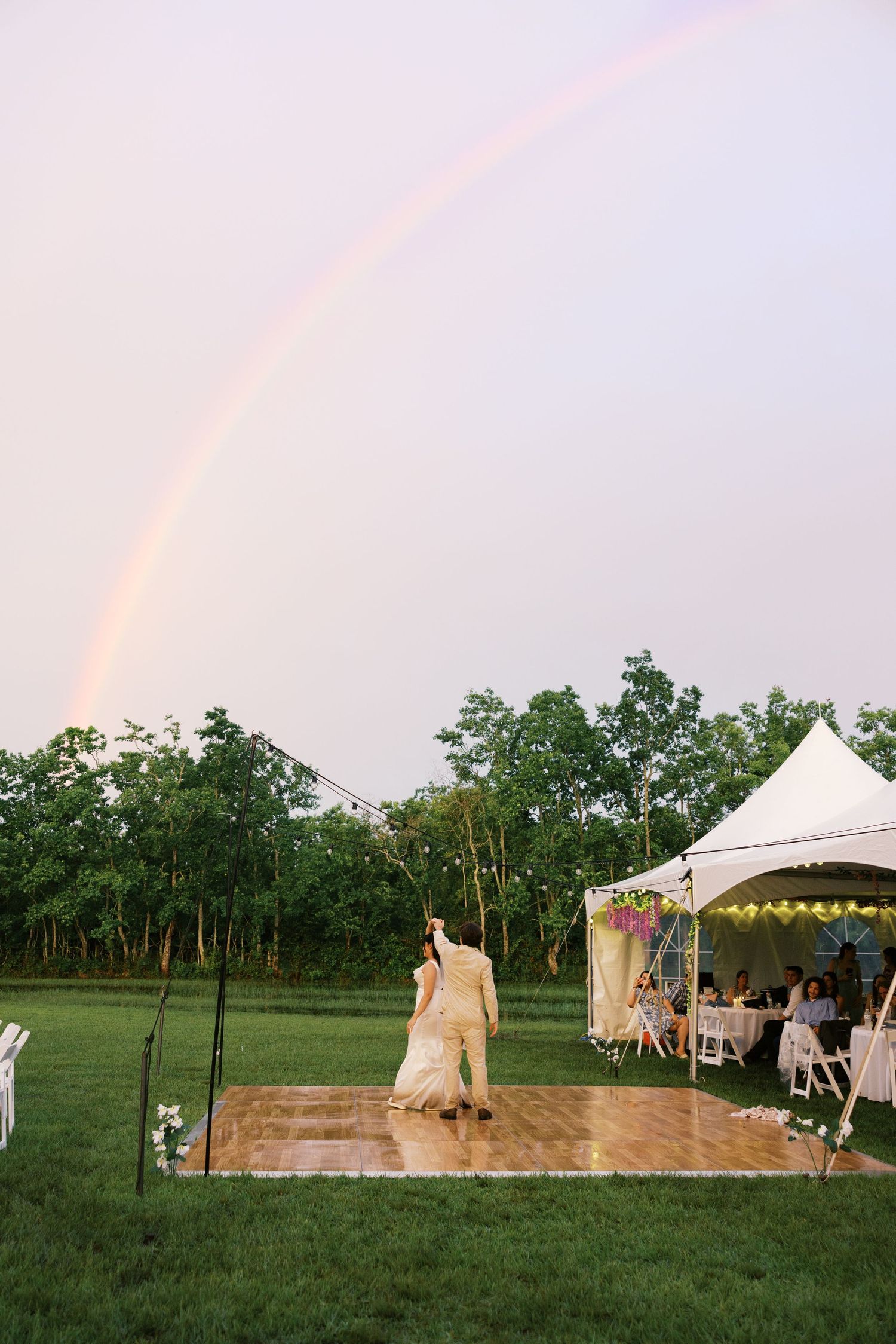 Couple sharing first dance on outdoor wooden floor under a stunning double rainbow at a tented wedding reception.