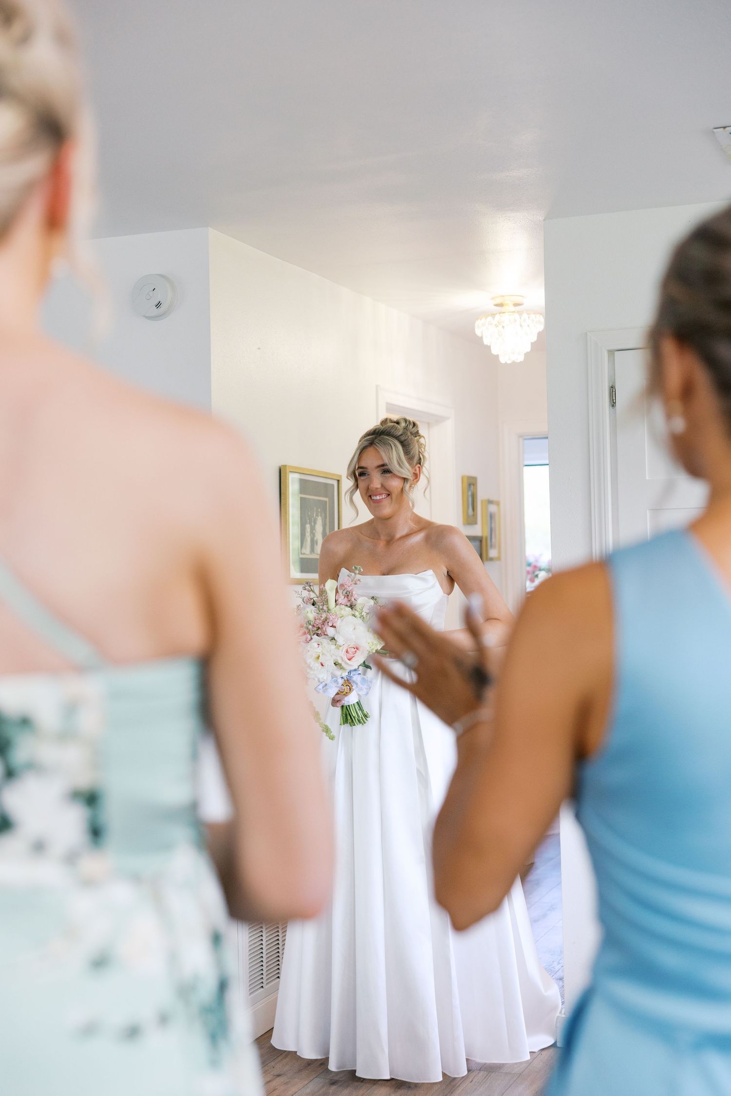 Smiling bride in white strapless gown holds pink bouquet while bridesmaids assist her getting ready.