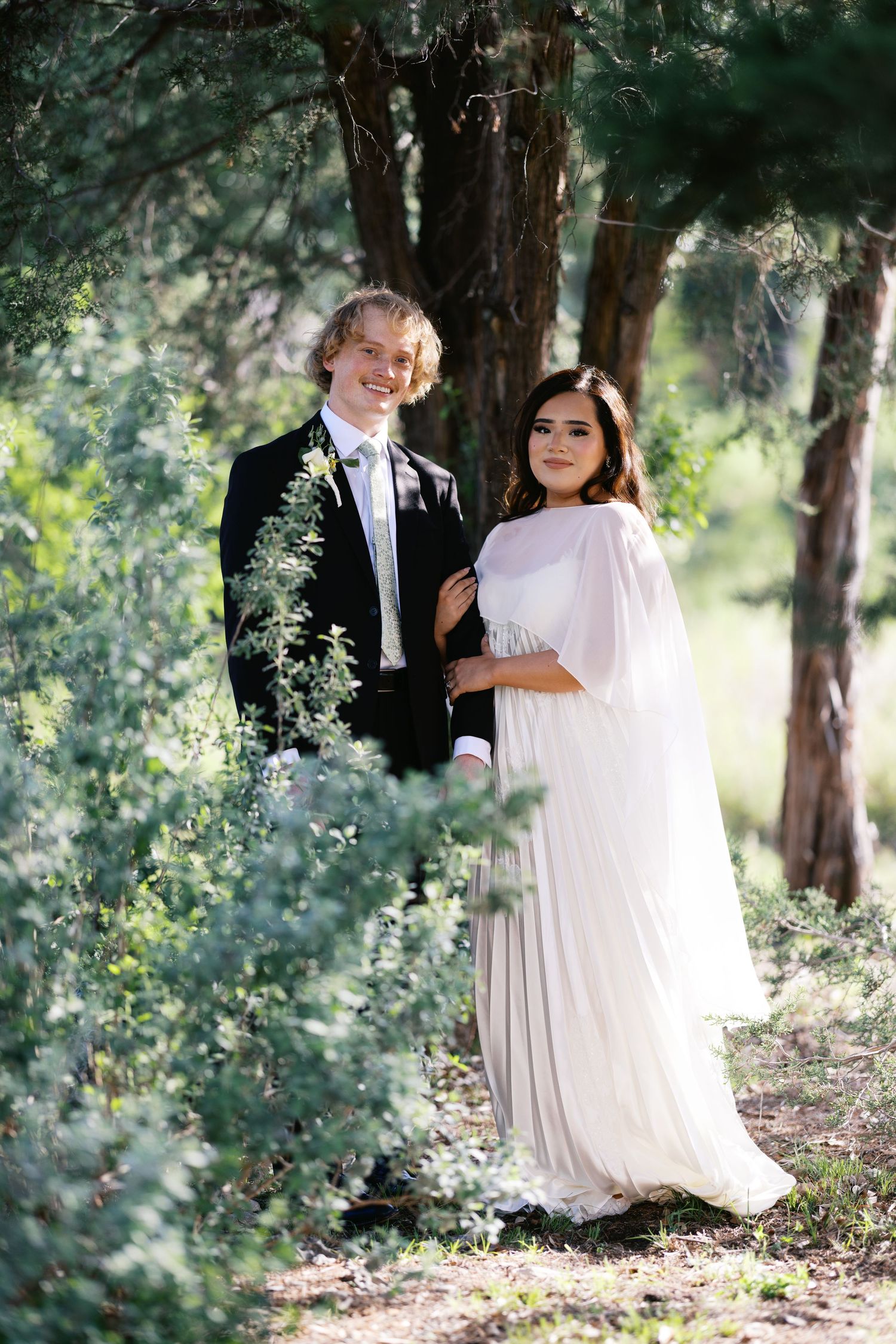 Bride and groom posing among lush green trees, bride in white cape gown, groom in black tuxedo.