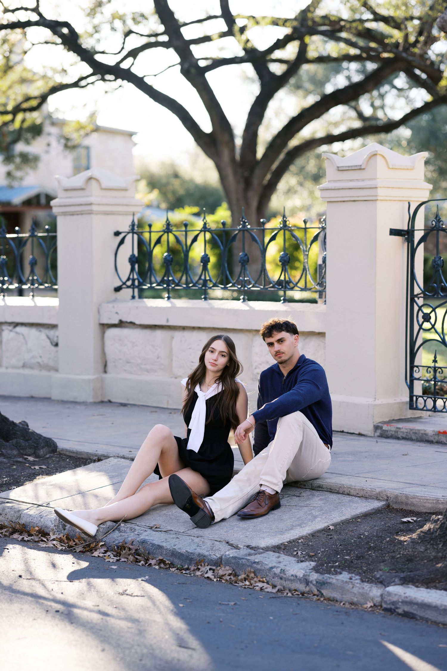 Young couple sitting together on steps near decorative iron fence under large oak tree outdoors.