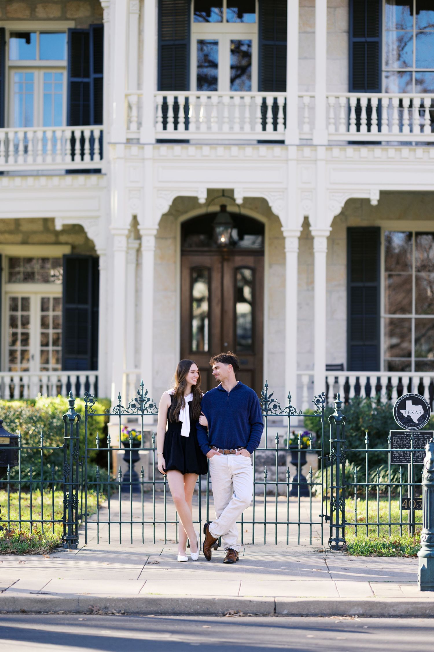 Couple walking arm in arm on a sidewalk in front of a grand white Southern mansion with iron fence.
