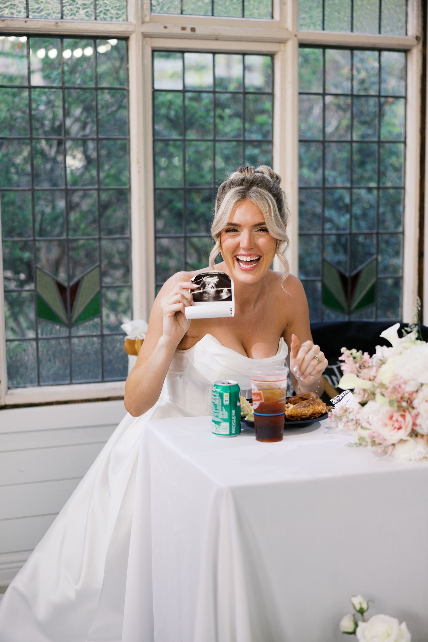Laughing bride in strapless gown holds ultrasound photo at wedding reception table.