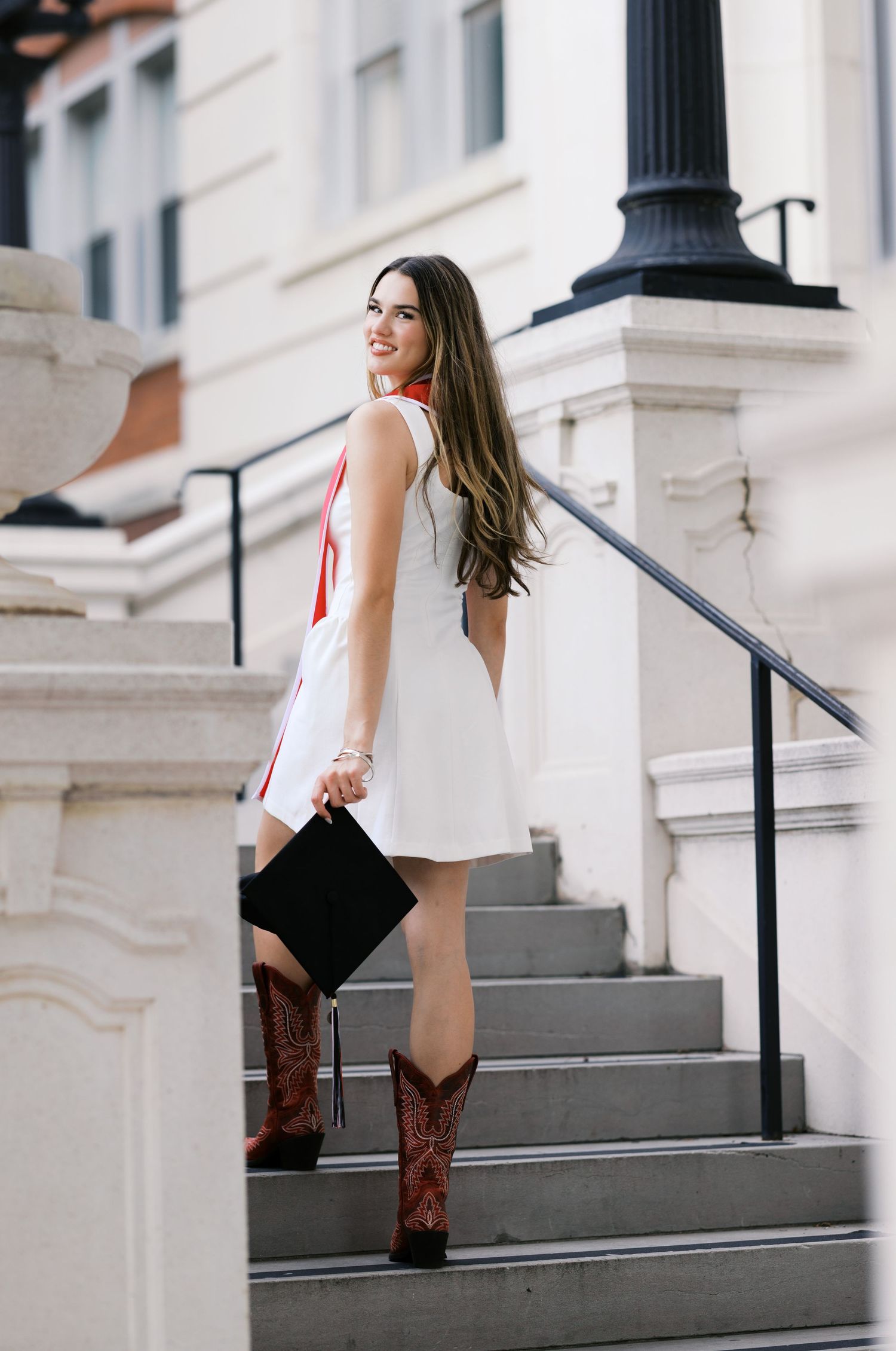 Young woman in white dress and cowboy boots holding graduation cap on outdoor steps.
