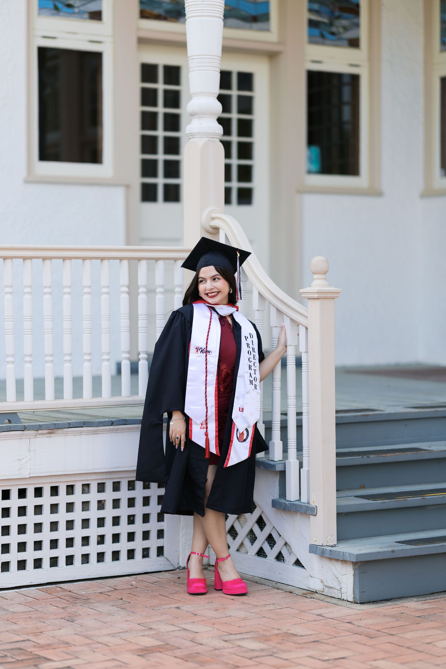 Young woman in black graduation gown, cap, and pink heels poses proudly on steps of a building.