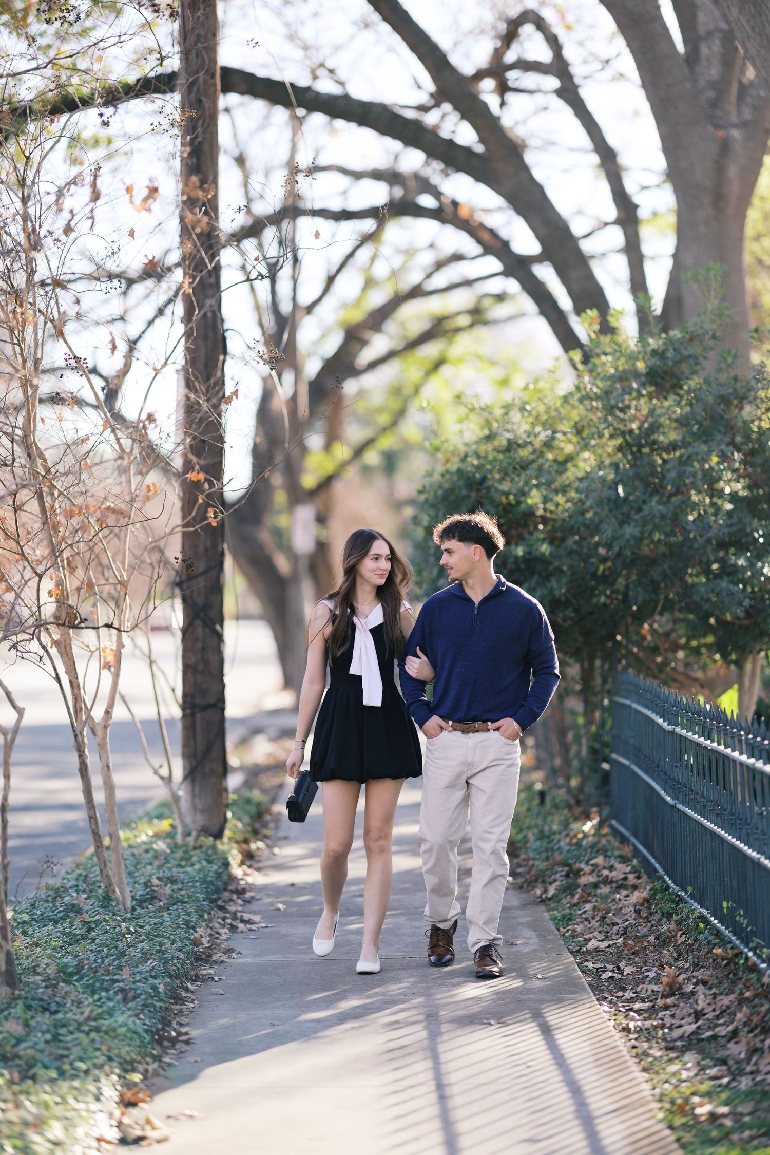 A couple walks hand-in-hand on a sunny sidewalk lined with trees, dressed in casual outfits, enjoying a leisurely stroll.