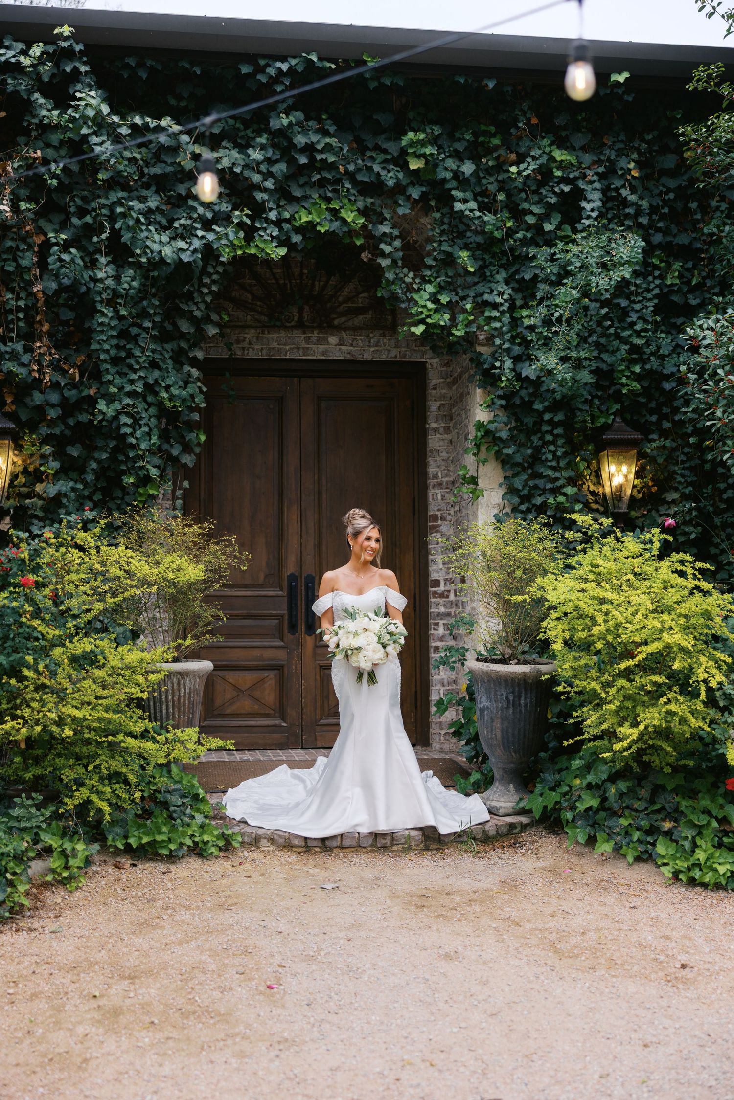 Bride in mermaid wedding dress poses before ivy-draped brick entrance with white bouquet.
