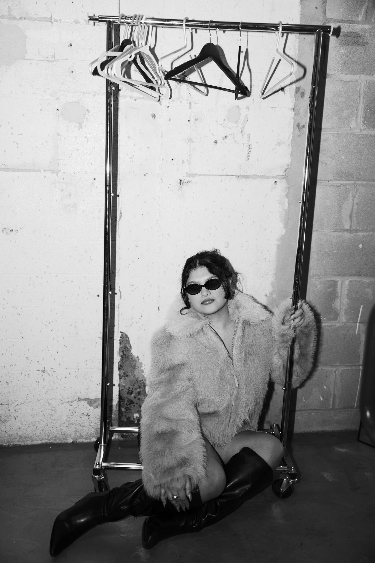 Artistic black and white photo of a figure in sunglasses and fur coat posing near clothing rack against brick wall.