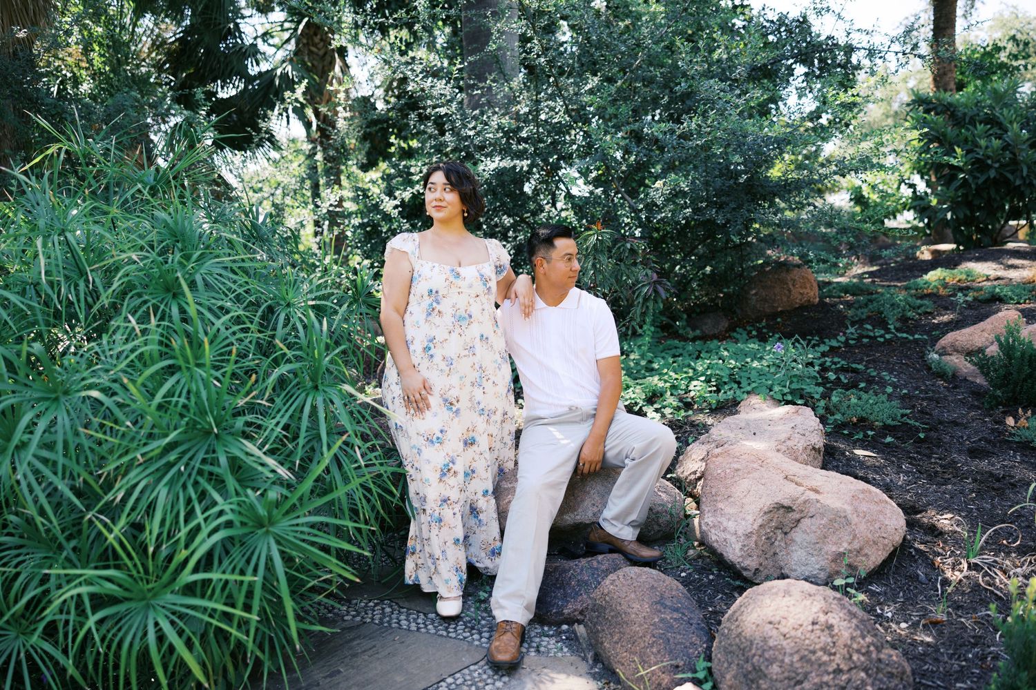A couple sits together on rocks in a peaceful garden setting with tropical plants.