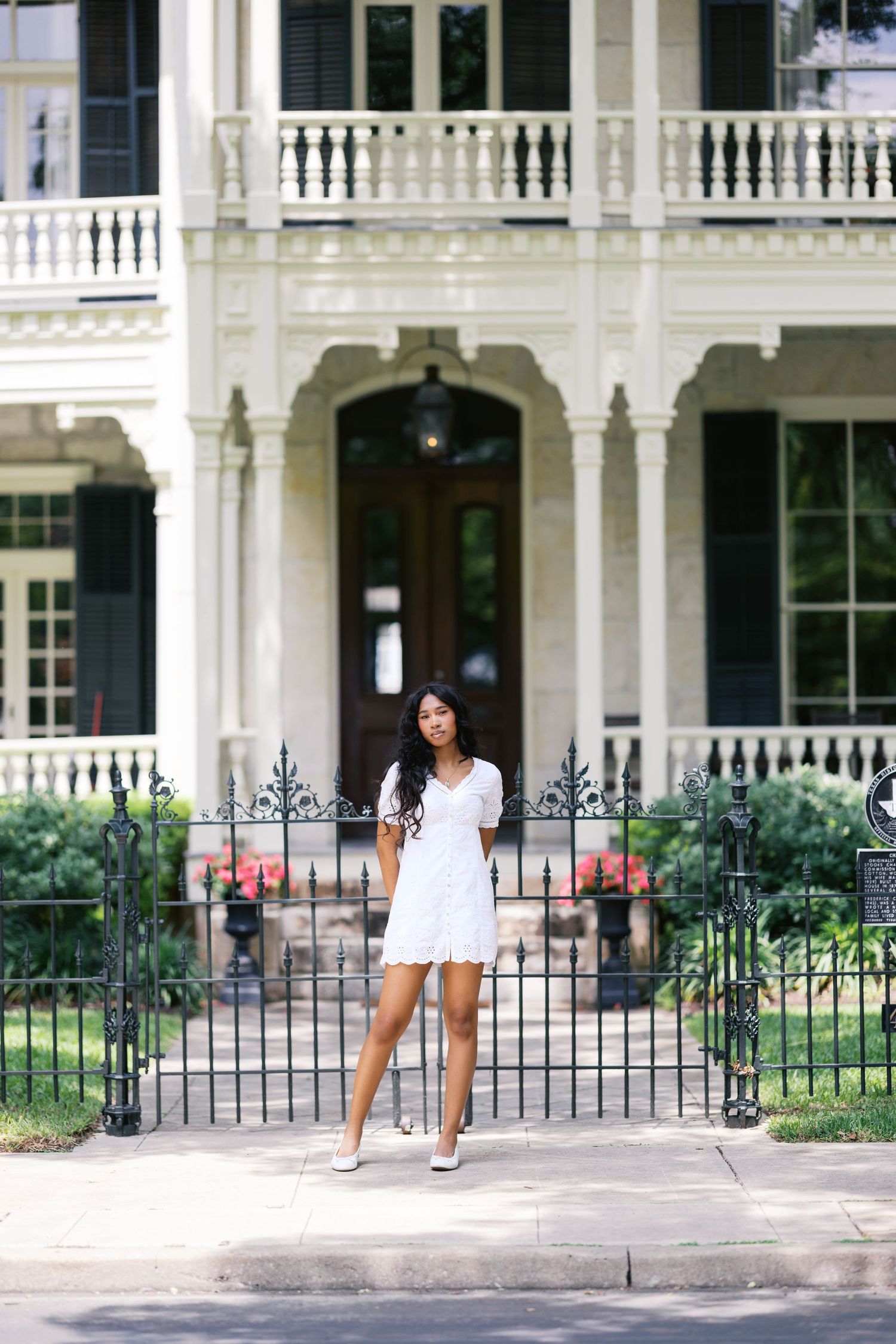 Elegant young woman in white dress poses before grand Victorian home with wrought iron fence and balcony.