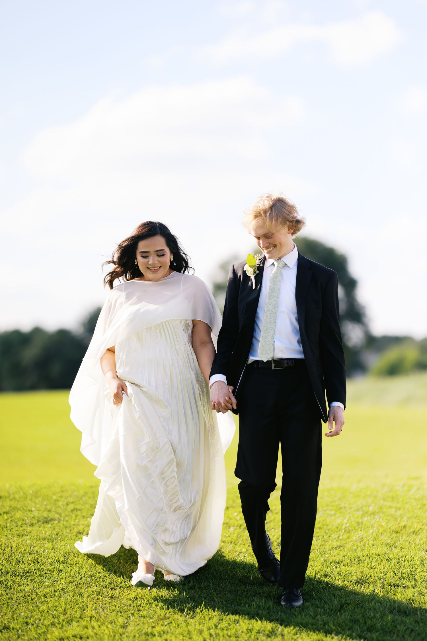 A bride and groom walk hand-in-hand across a lush green field on their wedding day.