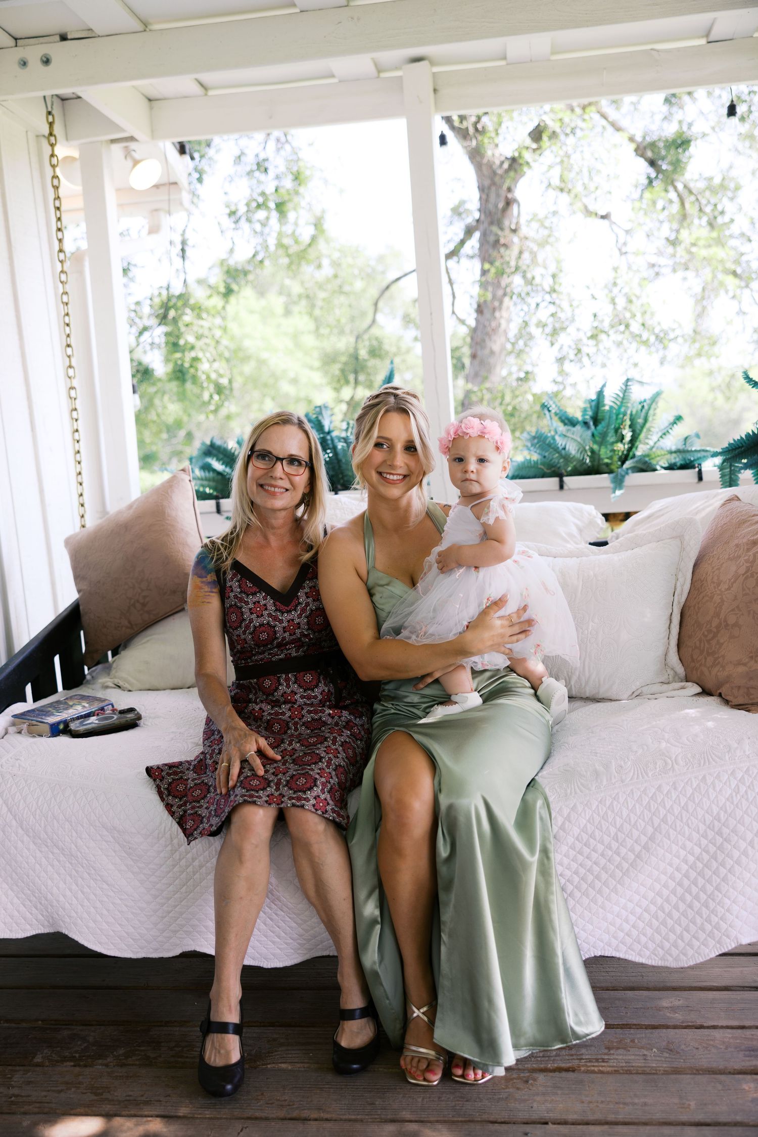 Two women and a baby in a floral headband pose together smiling on a porch daybed.