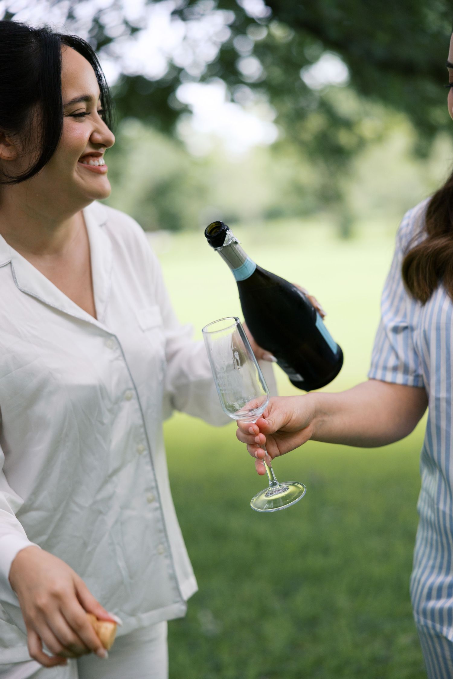 Woman in white pajamas smiling as champagne is poured into her glass outdoors at a wedding celebration.
