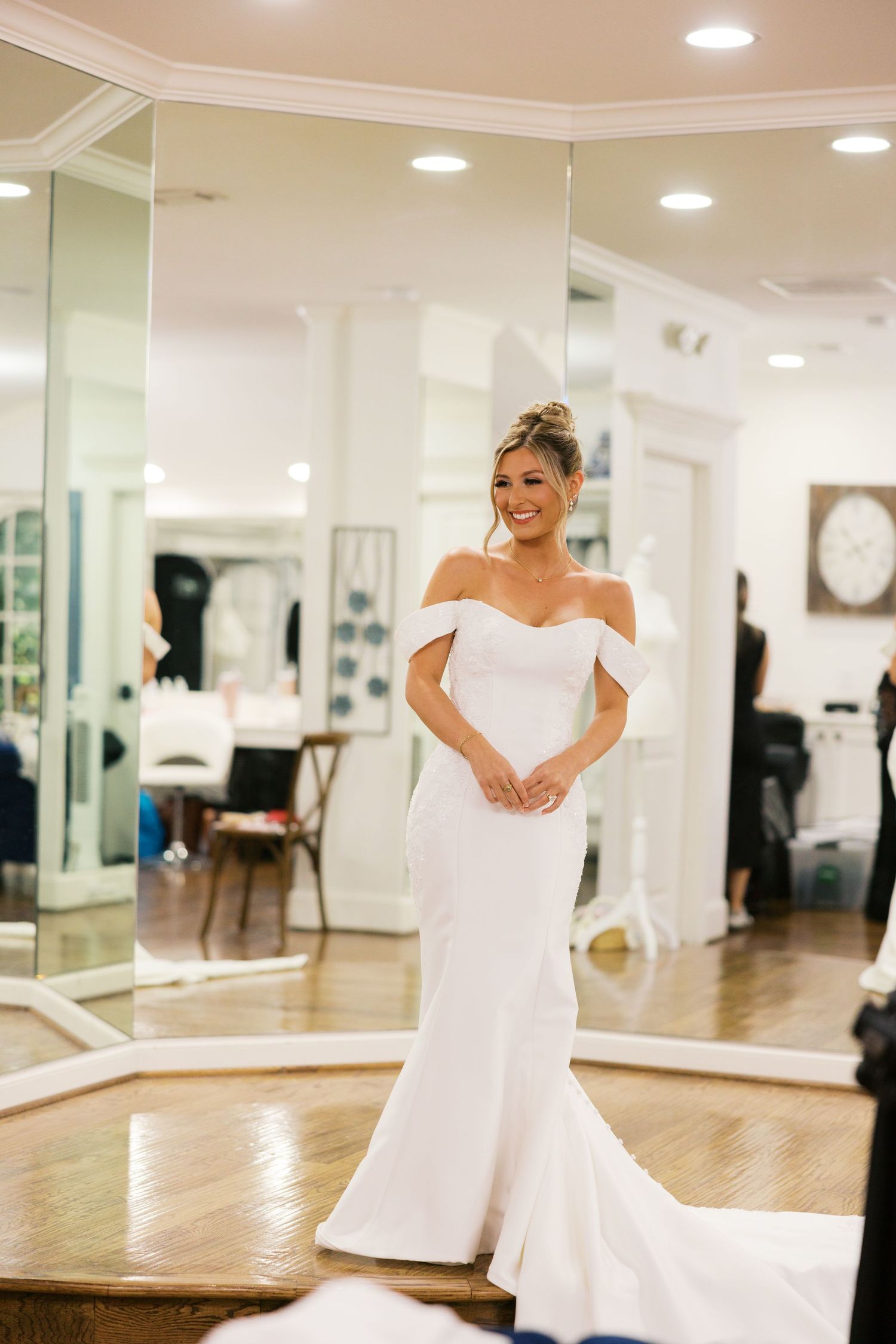 Smiling bride wearing elegant white off-shoulder mermaid wedding gown in a bridal boutique mirror room.