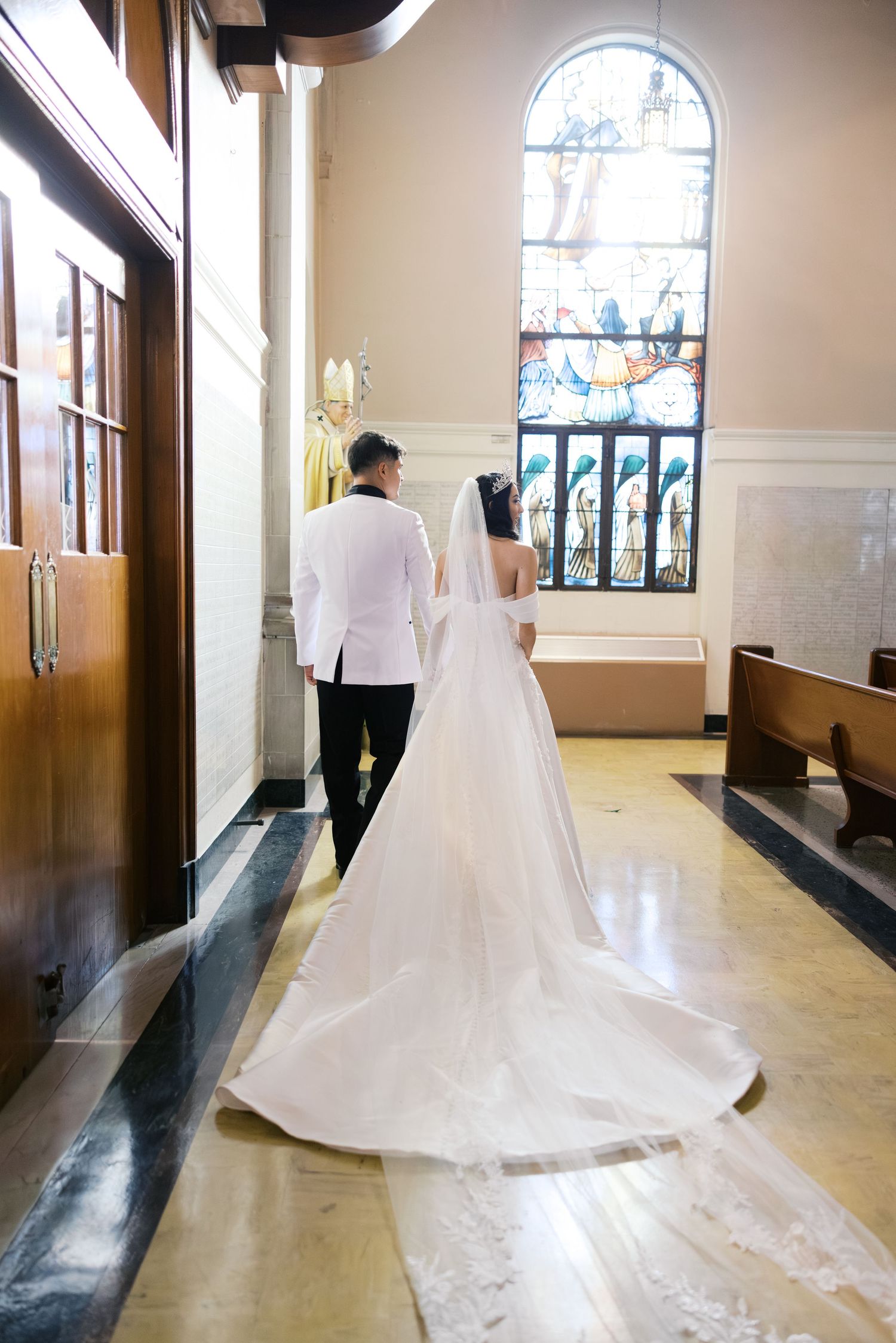 A couple in wedding attire walking down a church hallway with stained glass windows.