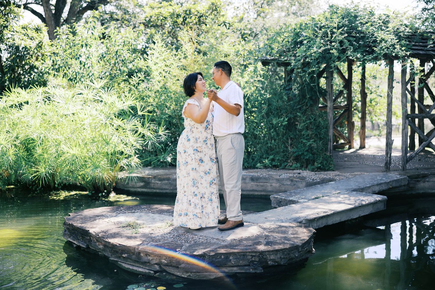 A couple shares an intimate moment while standing on a stone platform over a peaceful pond.
