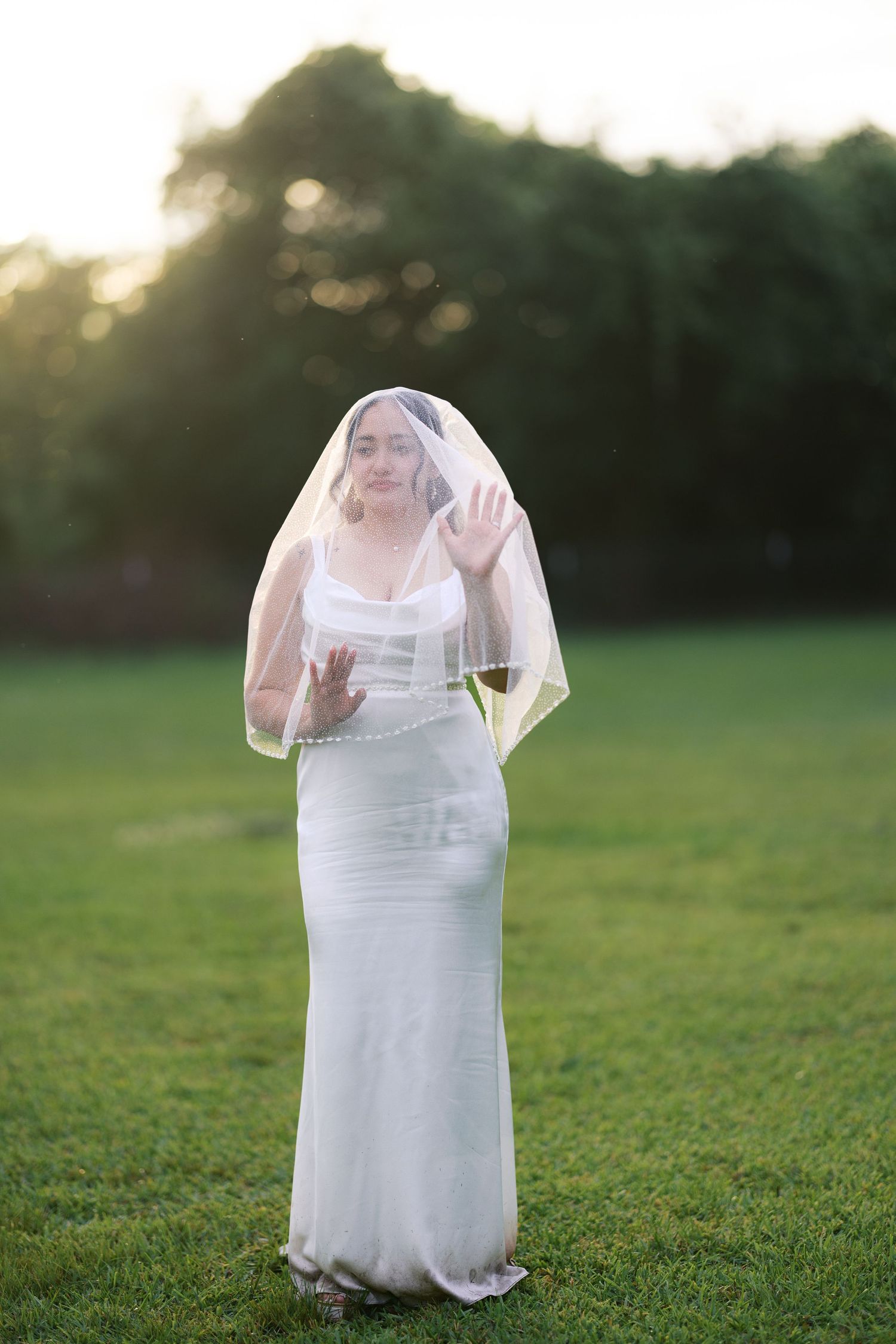Bride in fitted white gown and pearl-trimmed veil standing on green lawn at golden hour sunset.