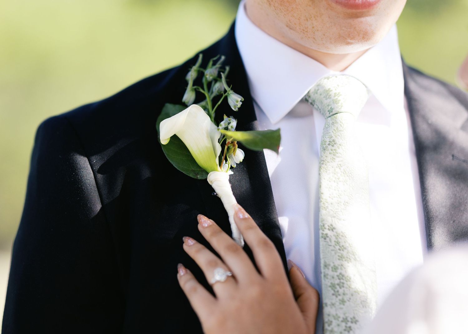 Groom in black suit with white calla lily boutonniere and sage green floral tie, bride's hand with ring on his chest.