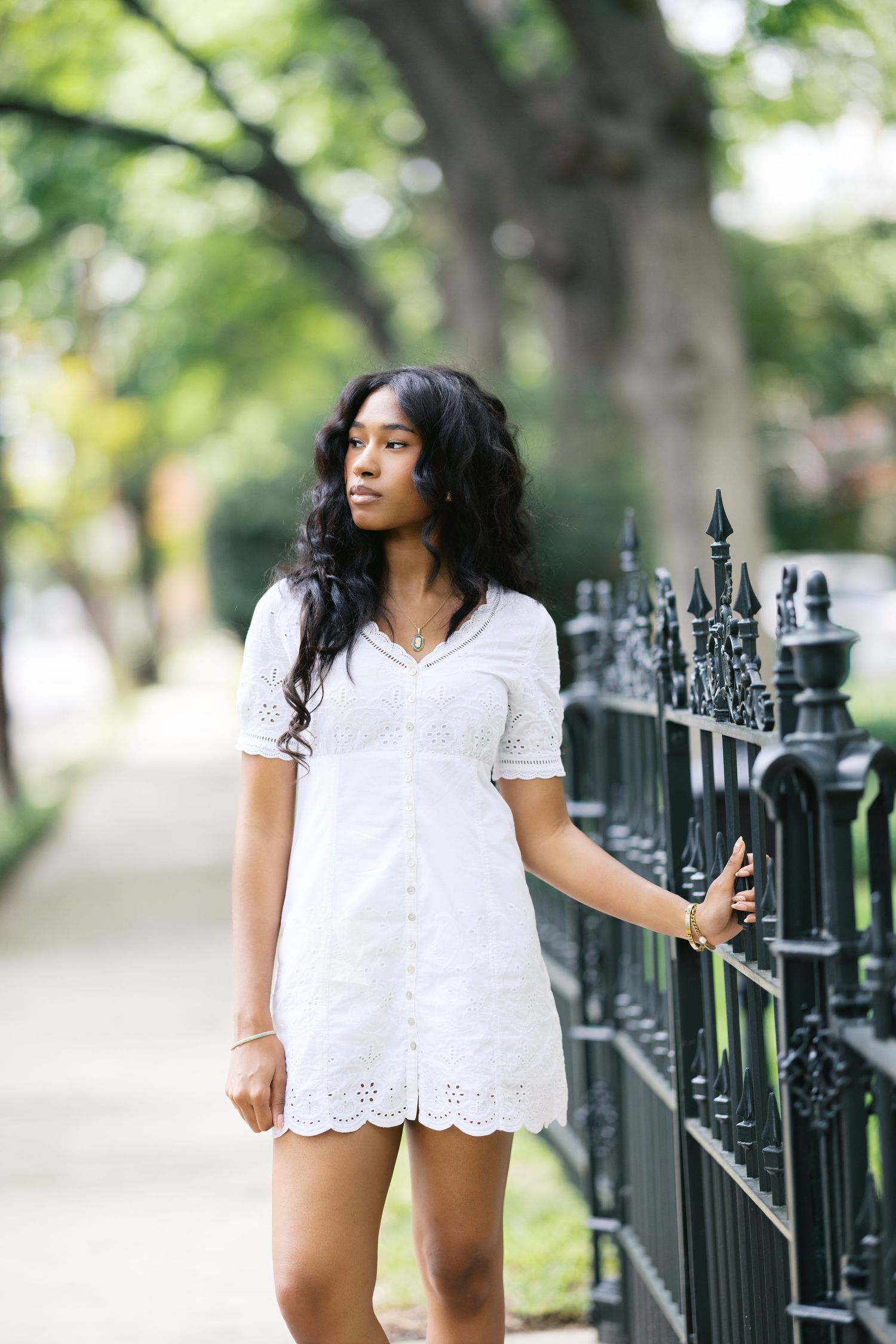 Young Black woman in white eyelet dress stands by ornate iron fence on tree-lined sidewalk.
