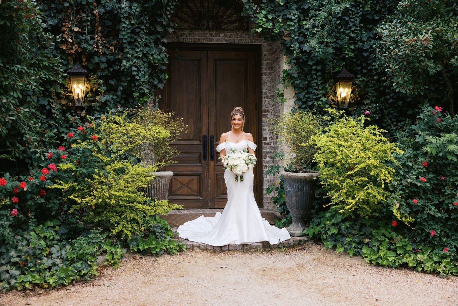 Bride in white mermaid gown holding bouquet stands before ivy-covered stone doorway with lanterns and lush greenery.