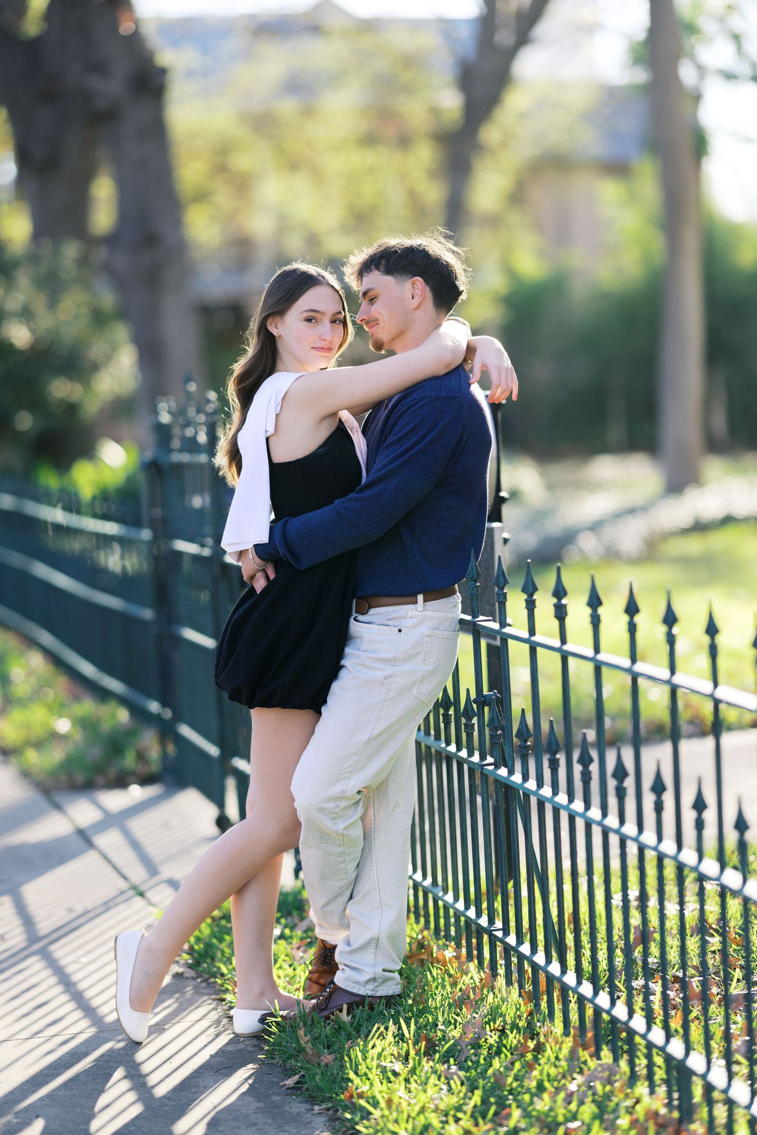 A romantic couple embraces by a green iron fence in a sunny park, dressed in black and navy outfits.