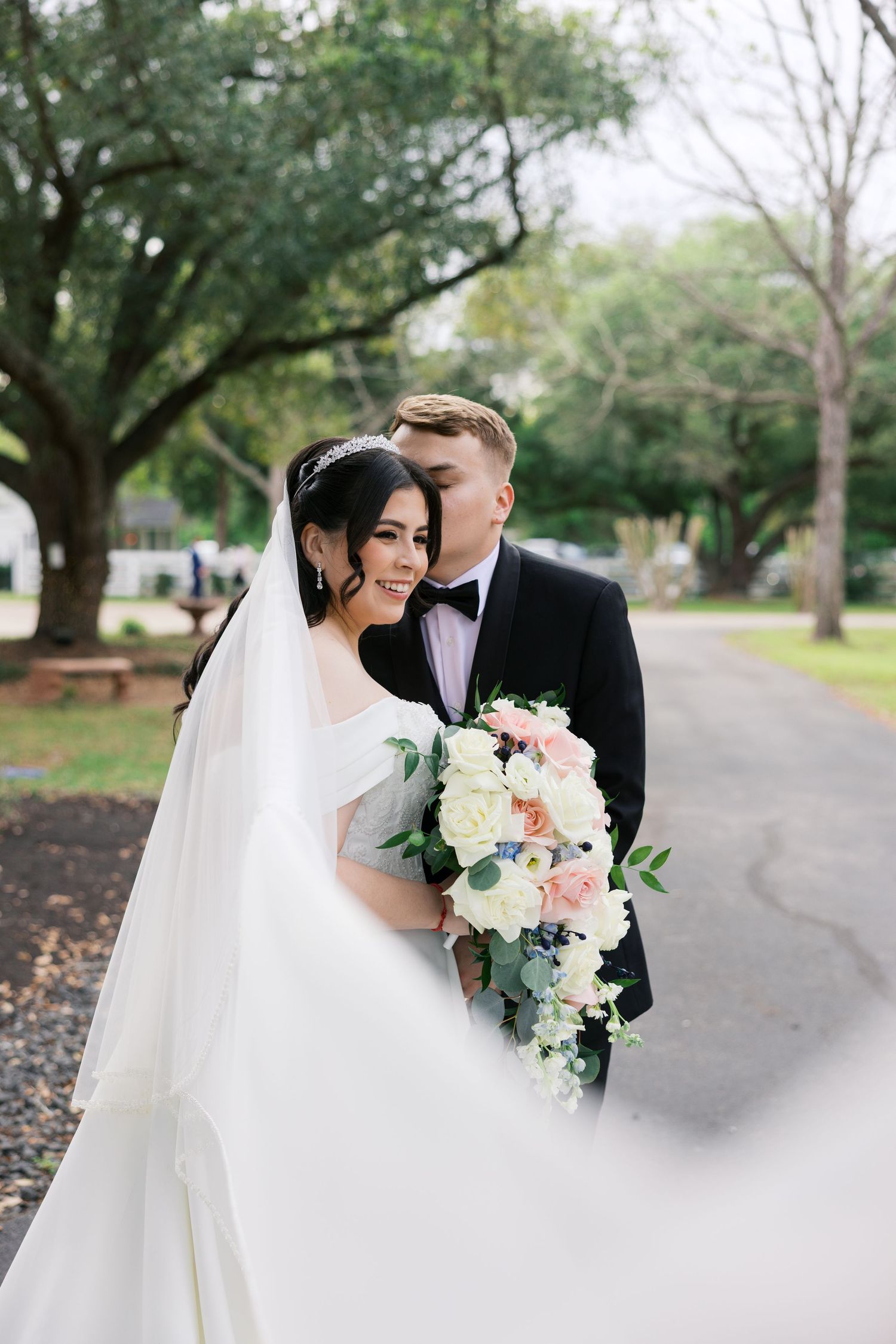 Bride and groom pose outdoors, she holds a pastel bouquet, veil flowing, as he tenderly kisses her cheek in a park.
