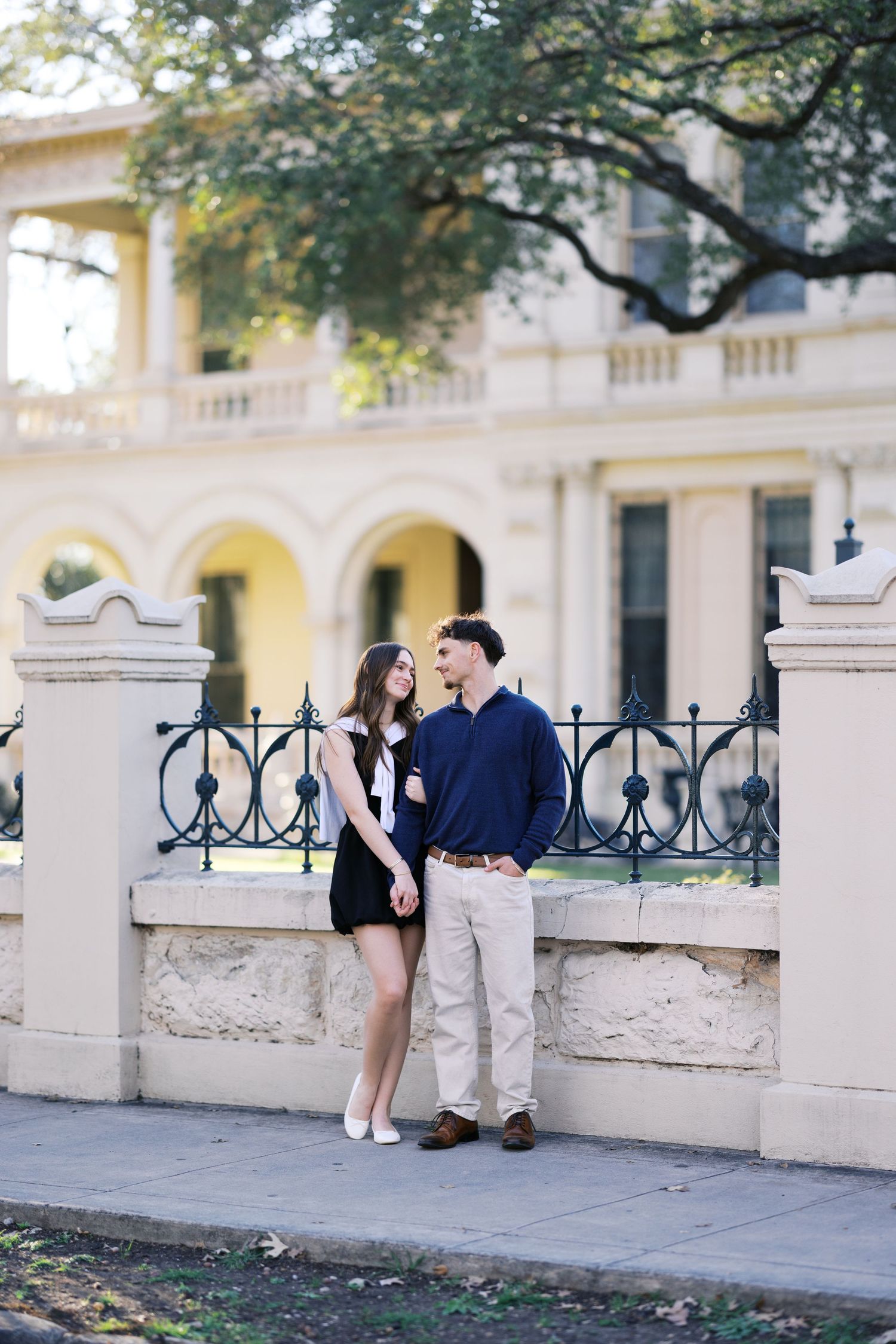Couple holding hands and gazing at each other by ornate iron fence outside elegant yellow historic building.