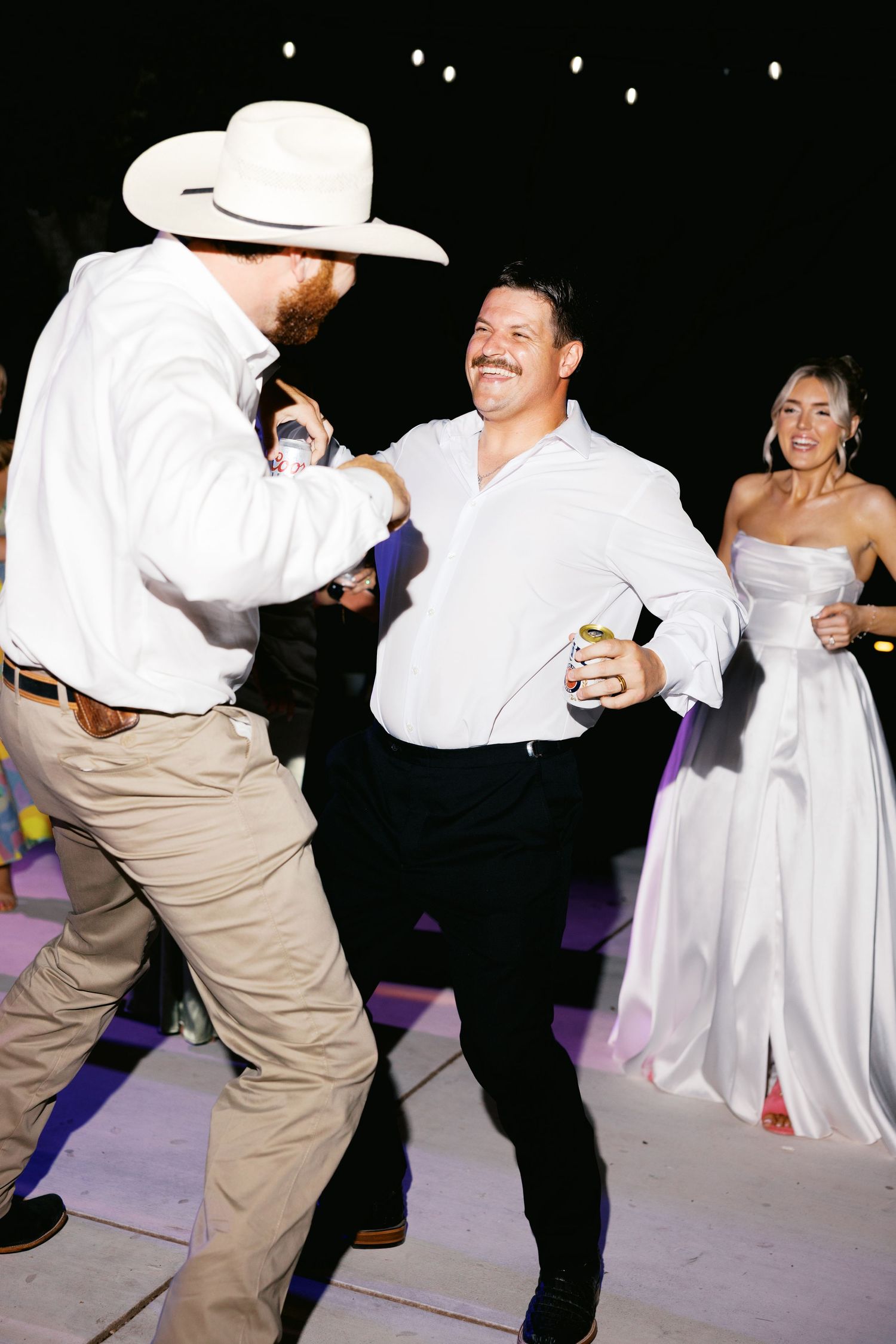 Groom dances joyfully with guest in cowboy hat at outdoor wedding reception at night.