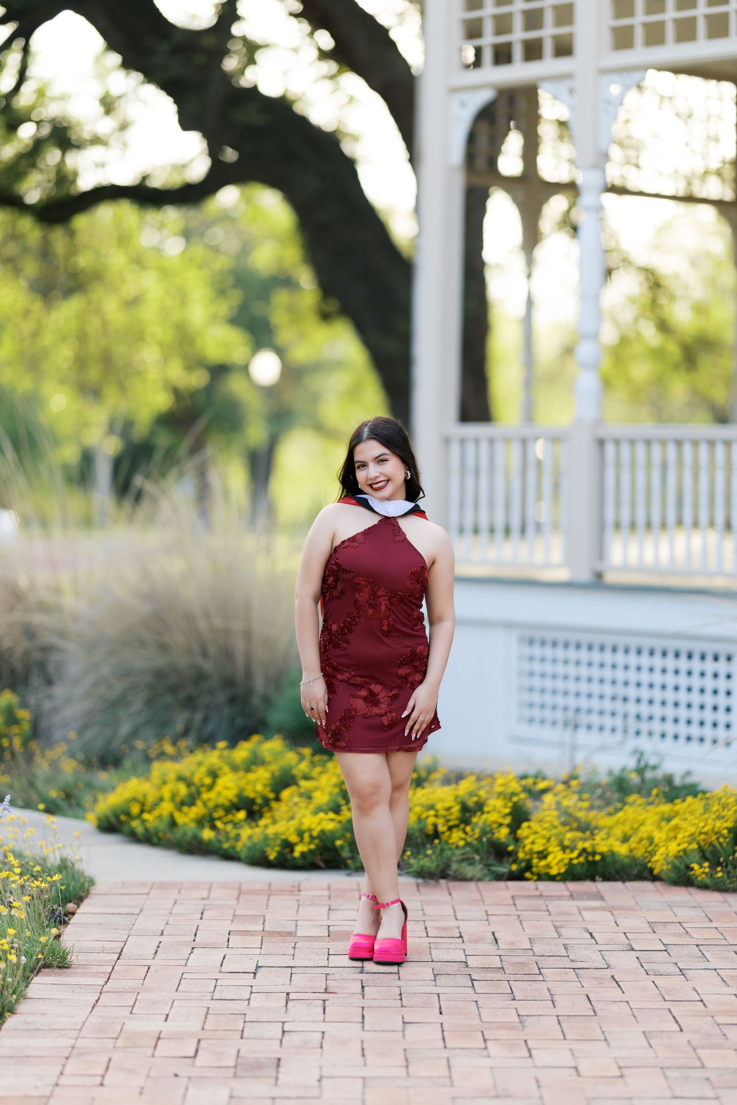 Young woman in burgundy halter dress and pink heels poses on brick path near white gazebo in garden.