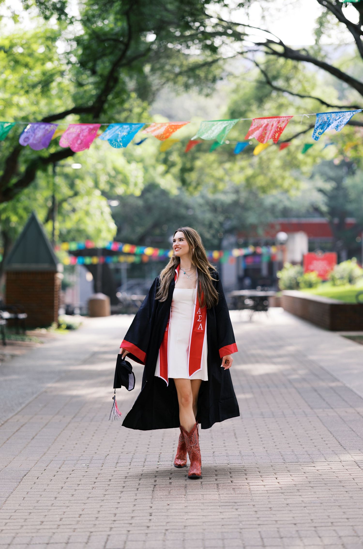 A smiling graduate walks in a tree-lined plaza wearing a black gown, red sash, and cowboy boots under colorful flags.
