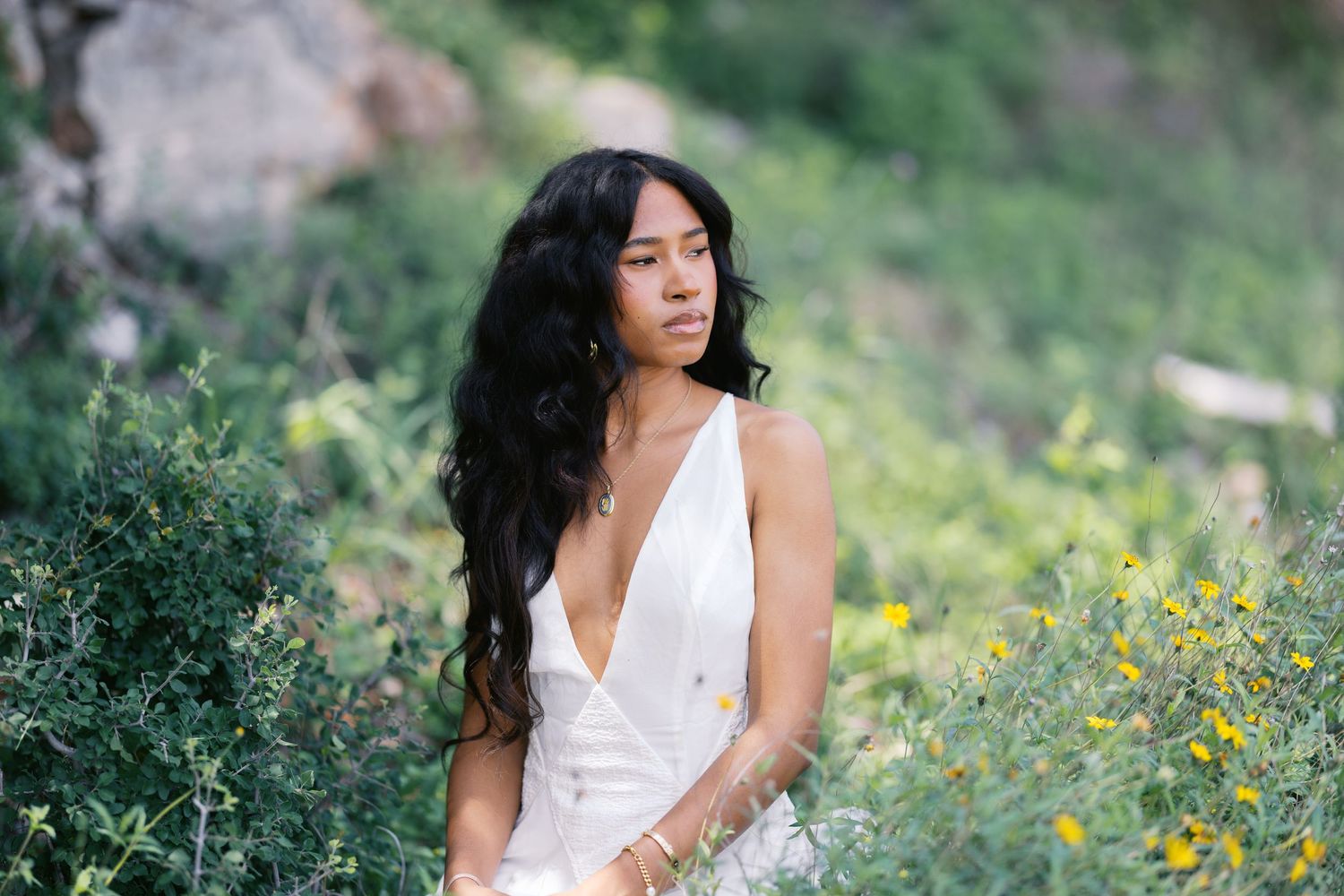 Young Black woman in white wrap dress sits among wildflowers and green shrubs in natural setting.